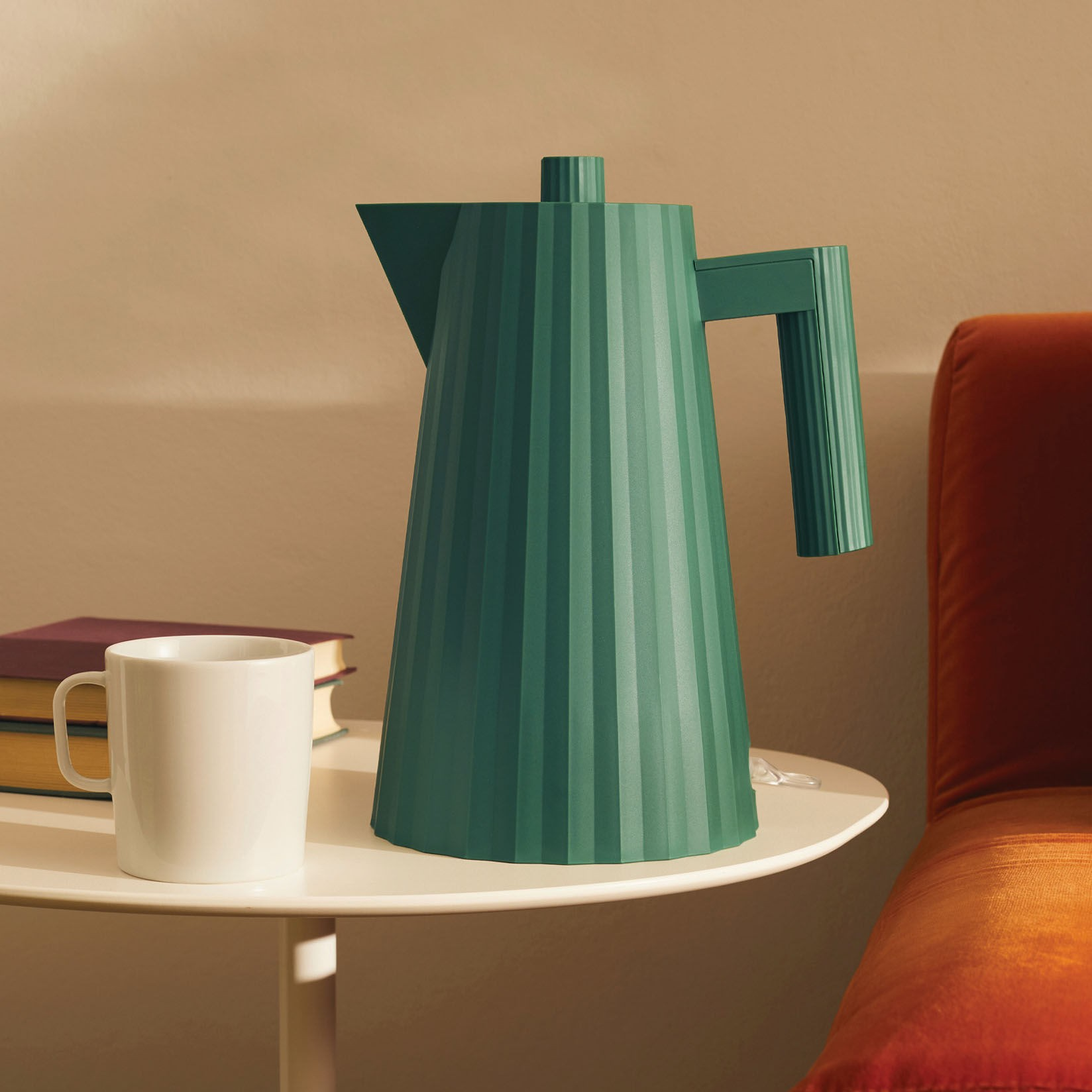 Bollitore elettrico verde su tavolo bianco, con tazza bianca, libri e sedia arancione sullo sfondo.