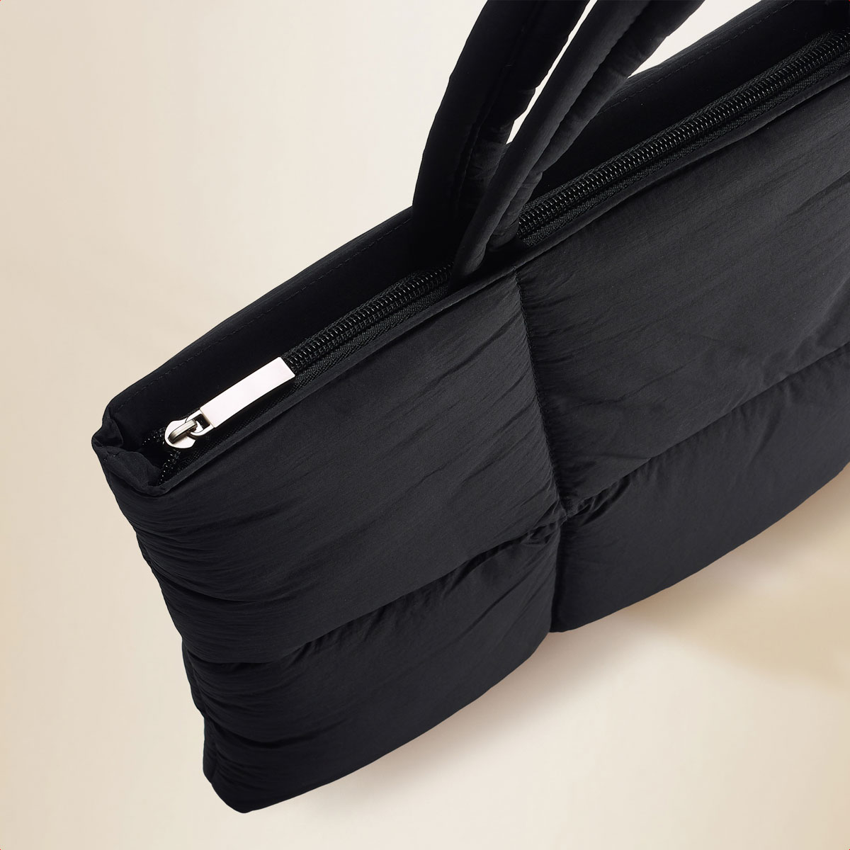 A black quilted tote bag, the bag is rectangular and has a zipper. The background is light beige.