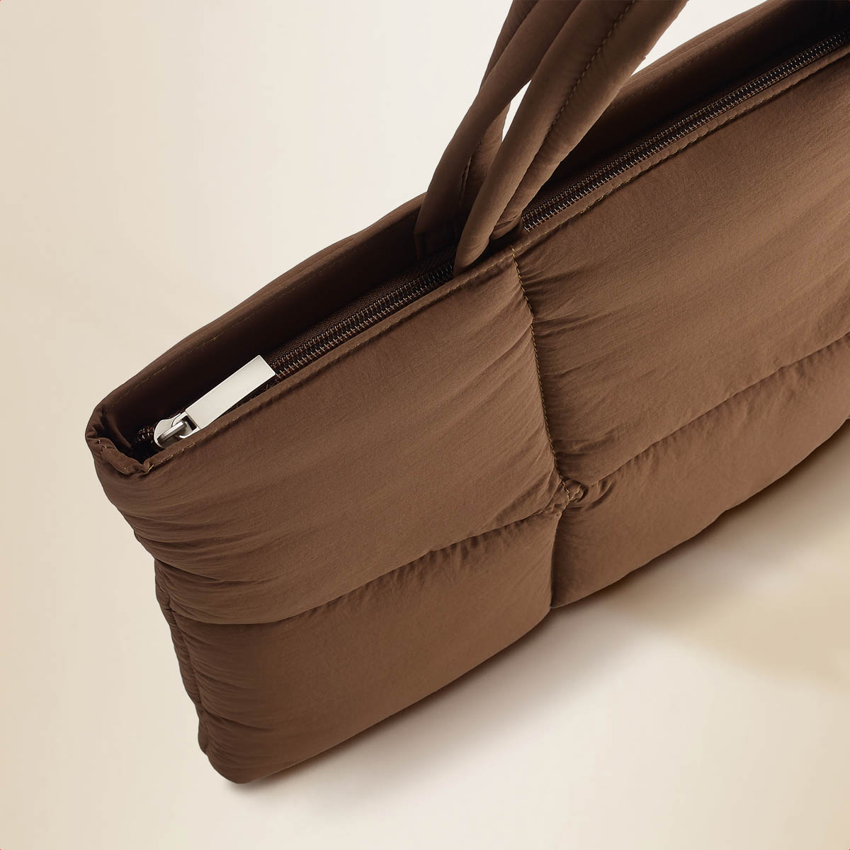 Brown quilted bag with zipper partially open. Handle visible. Shows texture and details of the stitching. Beige background.