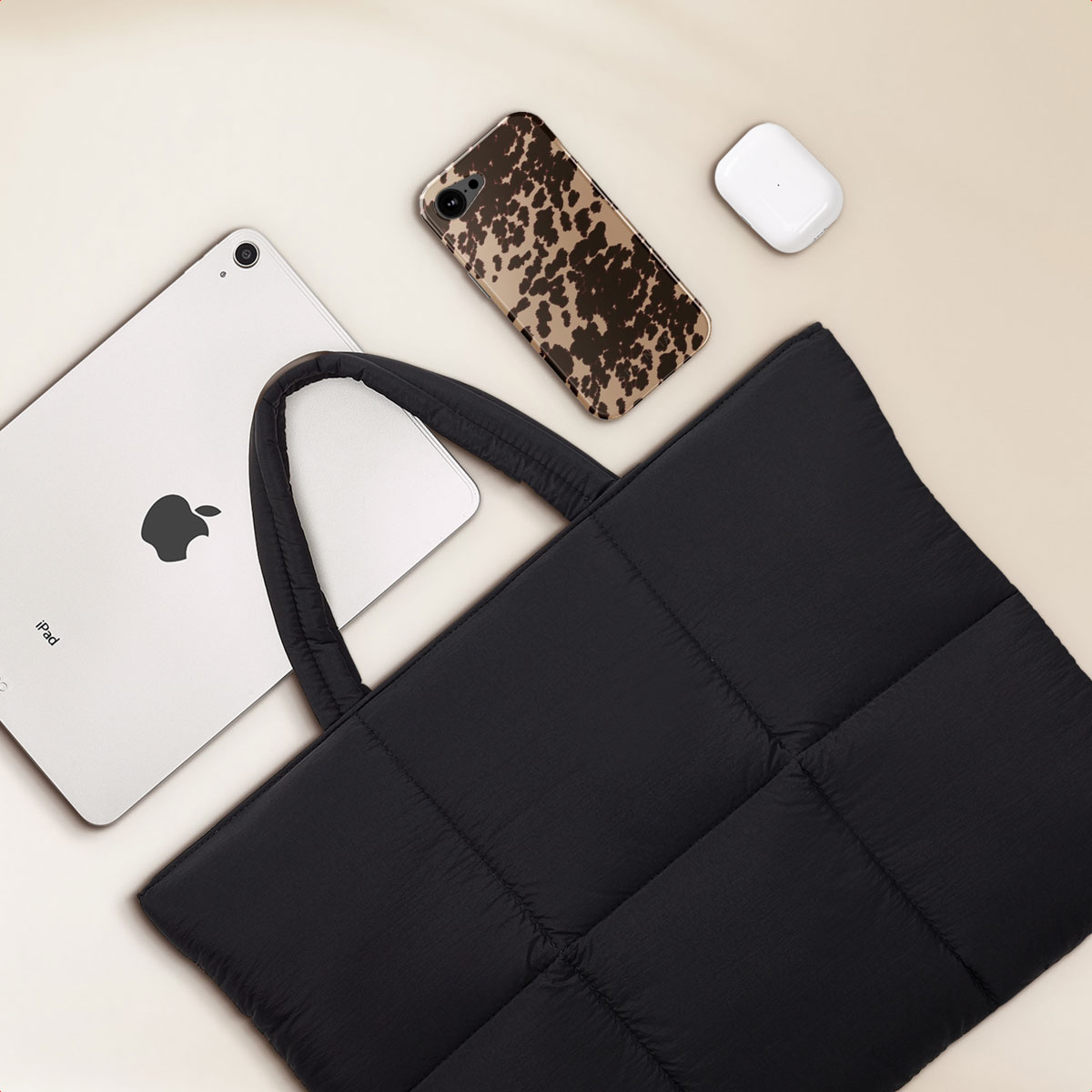 Black quilted tote bag with iPad, phone case, and earbuds on a light beige background.