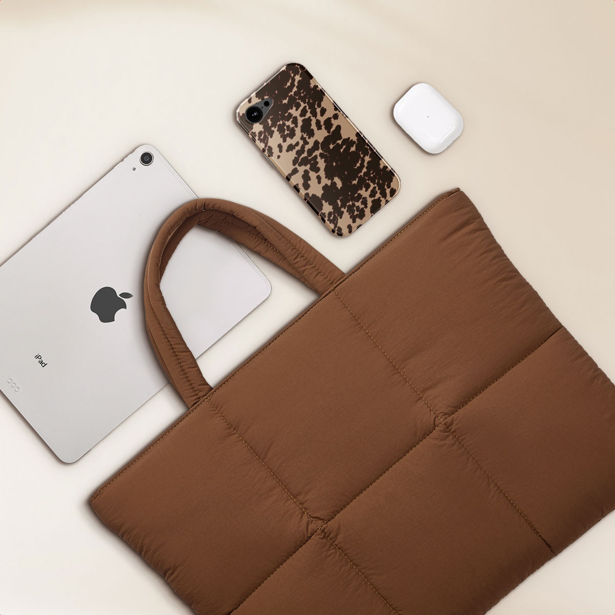 Brown quilted tote bag with iPad, phone case, and earbuds on a light beige background.