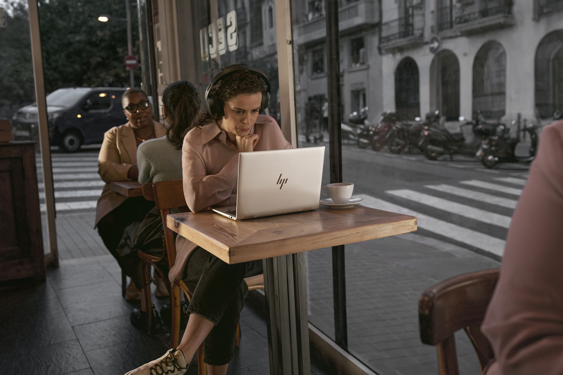 Mujer usando portátil en un café, con auriculares, otras personas y vista de la calle.