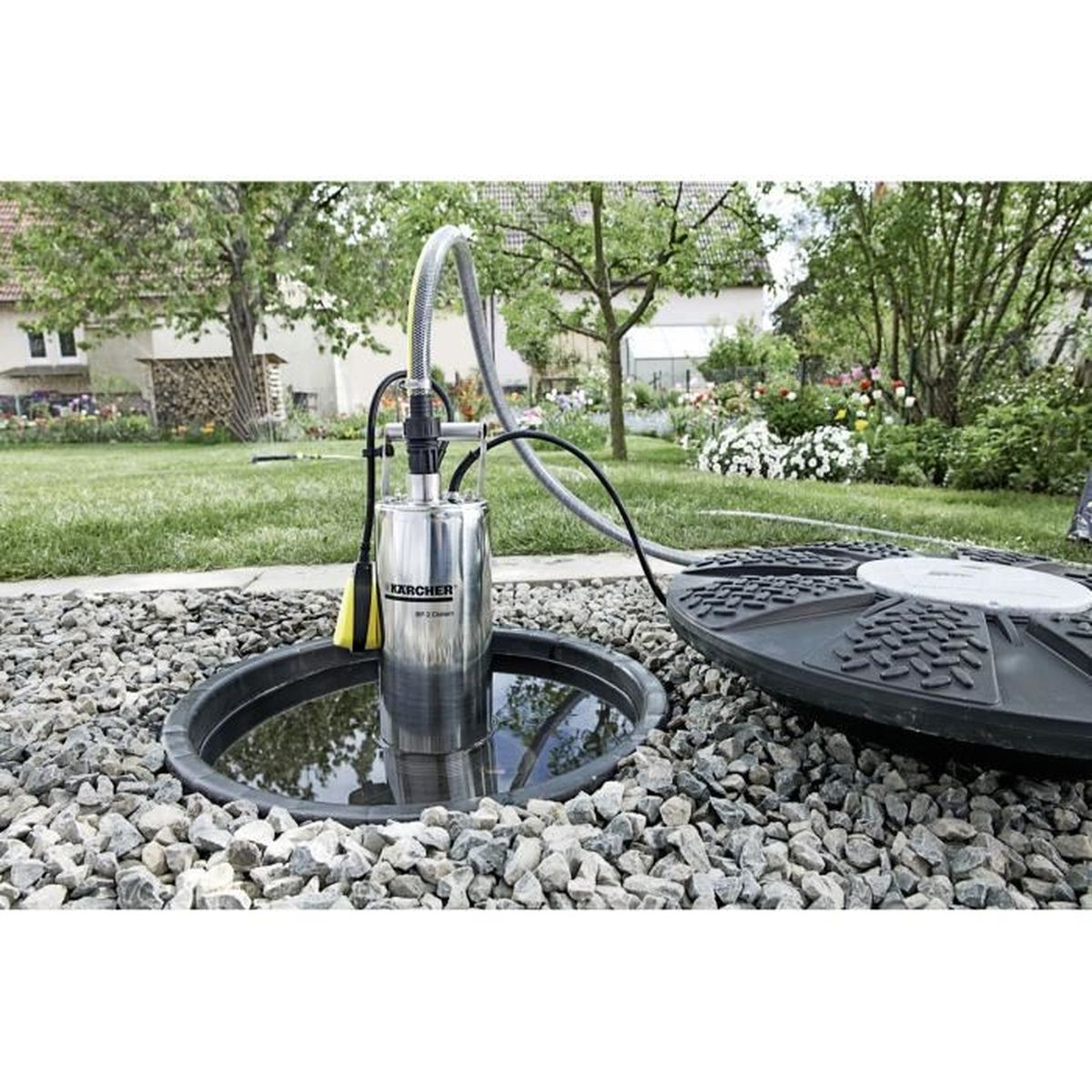 Eine Wasserpumpe in einem Steinbecken, angeschlossen an einen Schlauch. Die Szene spielt in einem Garten mit Bäumen und Gras.