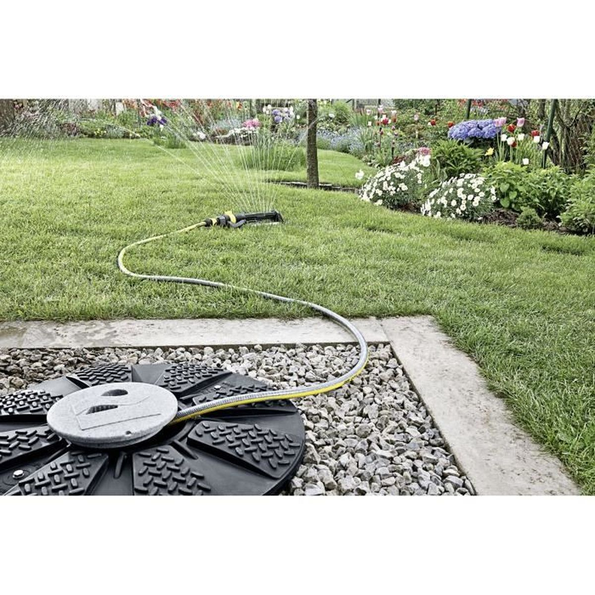 Ein Rasensprengersystem bewässert einen grünen Rasen. Ein Garten mit Blumen und einem Kiesbereich befindet sich im Hintergrund.