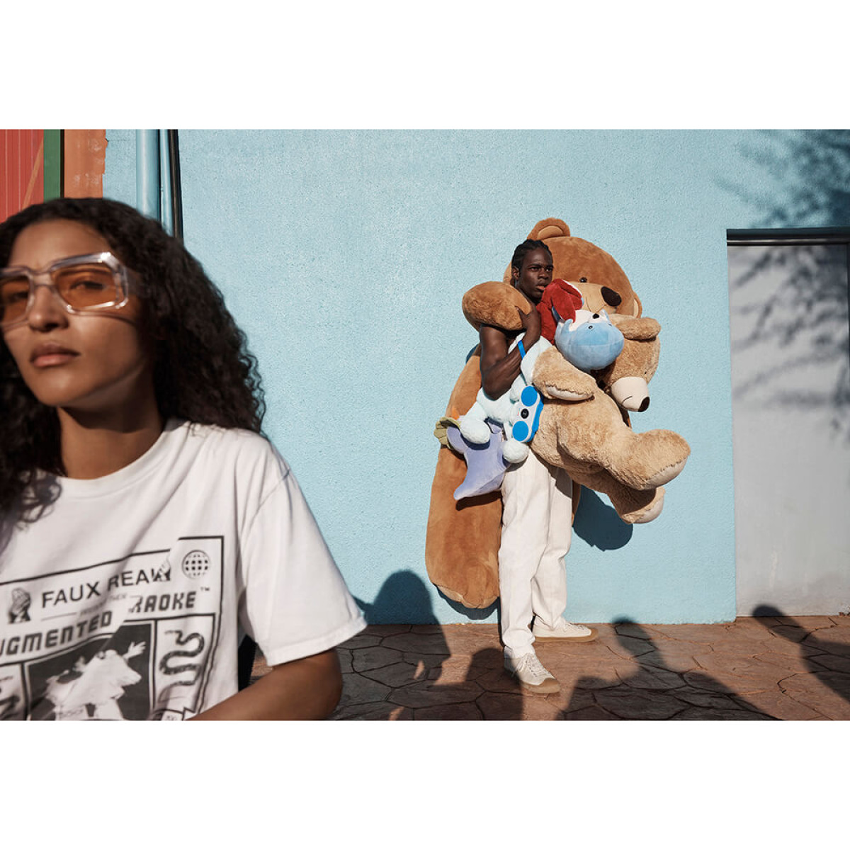 Osoba niesie dużego misia i pluszaki. Kobieta w okularach stoi po lewej. Niebieska ściana w tle.