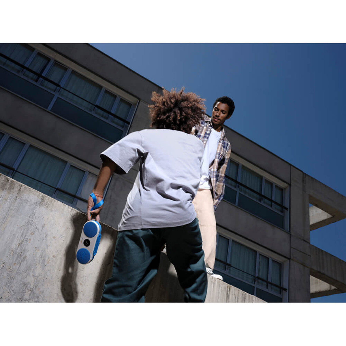 Dwie osoby stojące na murze. Jedna trzyma biało-niebieski głośnik. Tłem jest błękitne niebo.