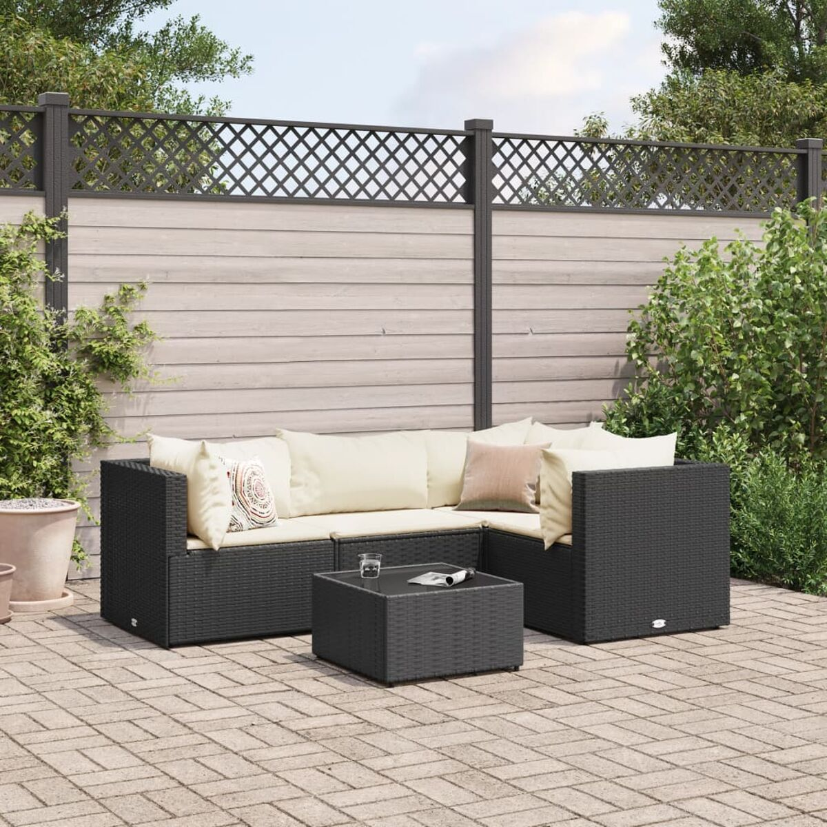 Conjunto exterior con sofá seccional de mimbre negro, mesa de centro cuadrada y patio de ladrillo. Arbustos verdes y valla gris.