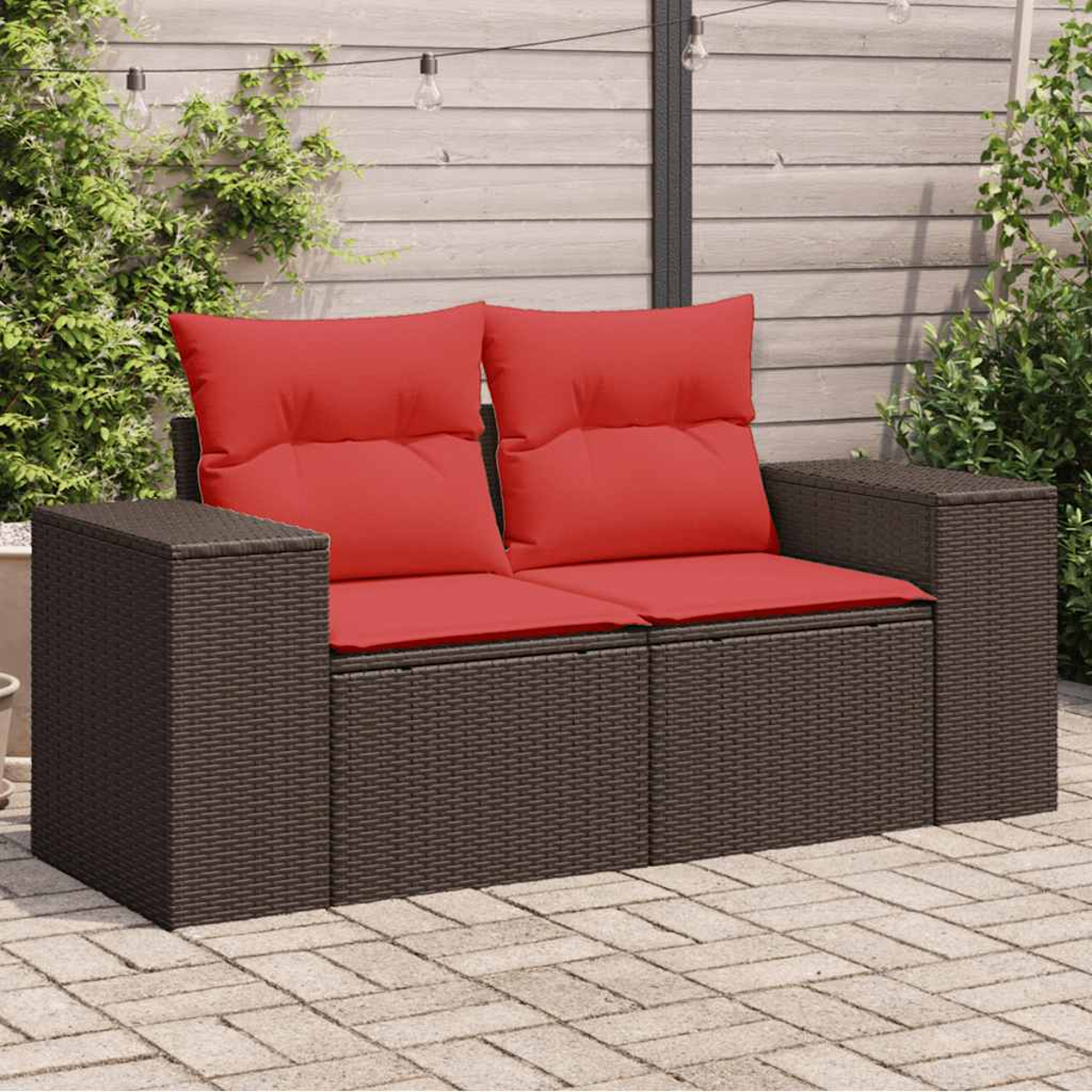 Outdoor-Korbsessel mit roten Kissen auf einer Ziegelterrasse, umgeben von Pflanzen. Sonniger Tag.