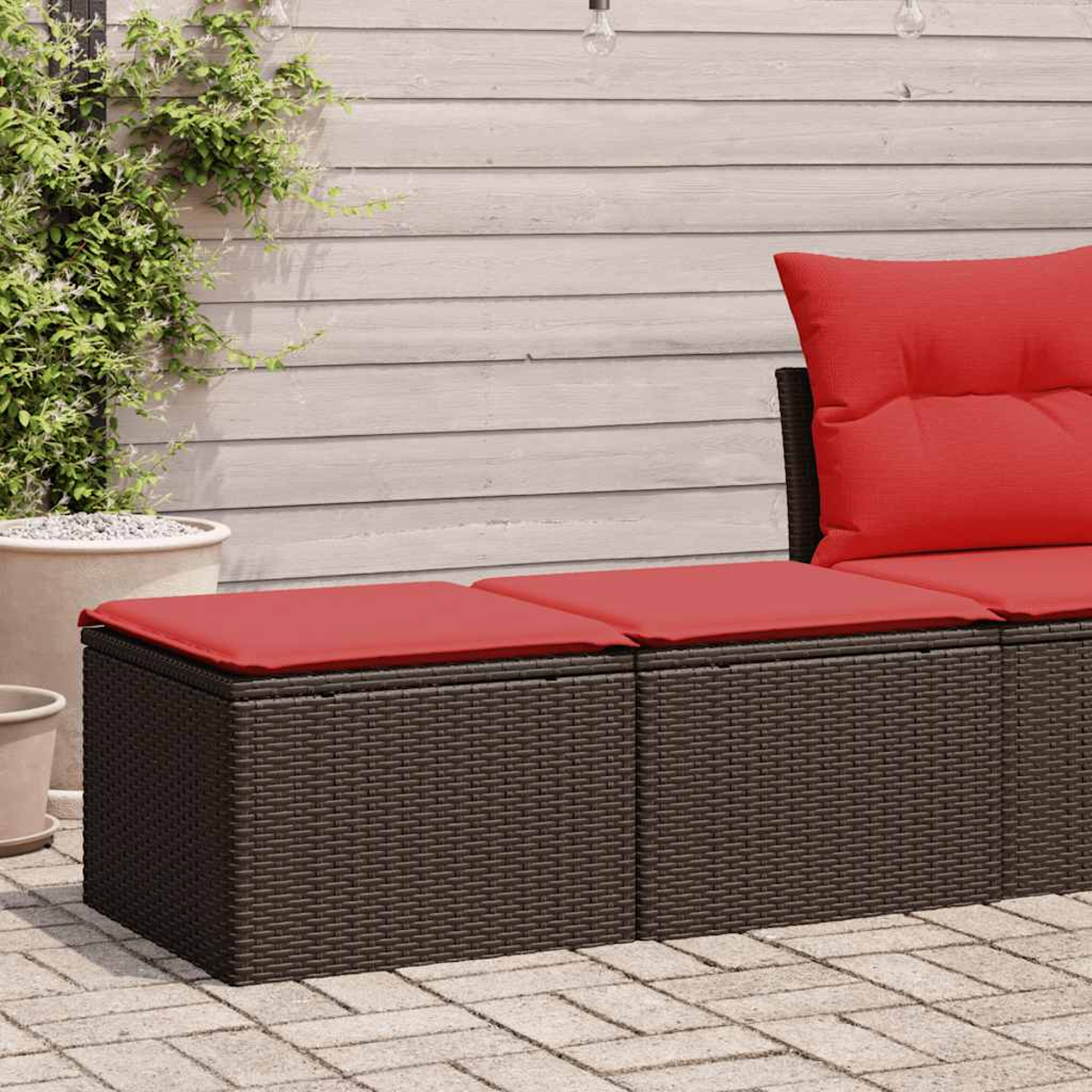 Nahaufnahme eines braunen Korbmöbels mit roten Kissen auf einer Ziegelterrasse. Eine kleine Pflanze ist zu sehen.