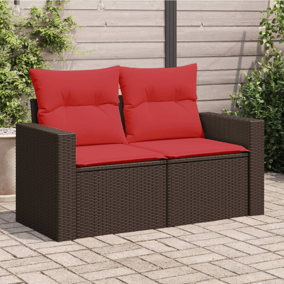 Ein braunes Korbsessel-Zweisitzer-Sofa mit roten Kissen auf einer Backstein-Terrasse. Pflanzen im Hintergrund.