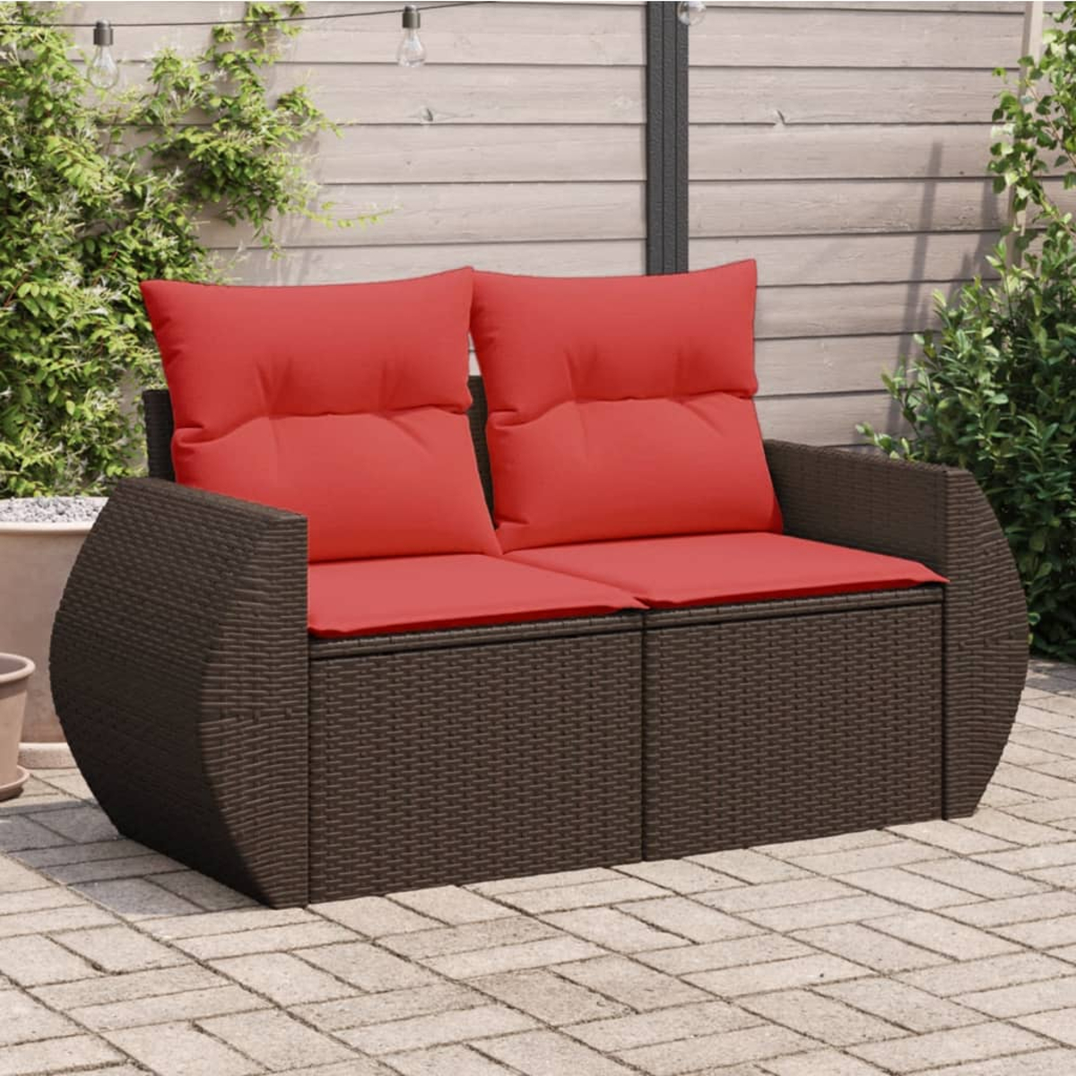 Braunes Rattan-Sofa mit roten Kissen auf Backstein-Terrasse. Hintergrund: Pflanzen, Holzverkleidung.