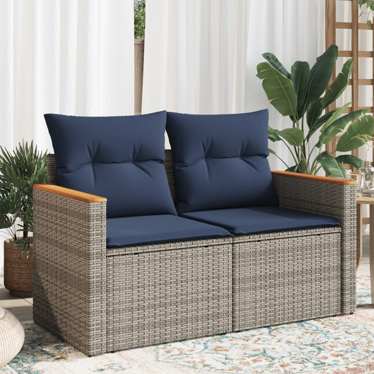 Graue Rattan-Loveseat mit blauen Kissen und Holzarmlehnen, auf einer Terrasse mit Pflanzen und Vorhängen.