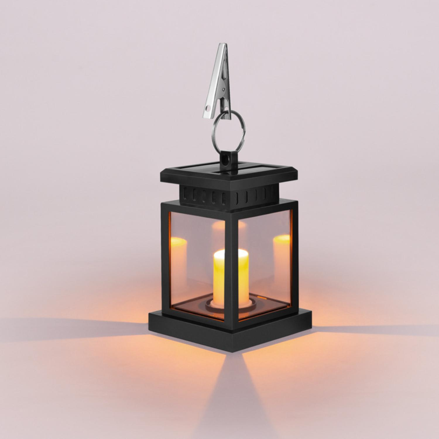 Eine schwarze Solar-Laterne mit kerzenähnlichem Licht, die an einem Metallclip vor rosa Hintergrund hängt.