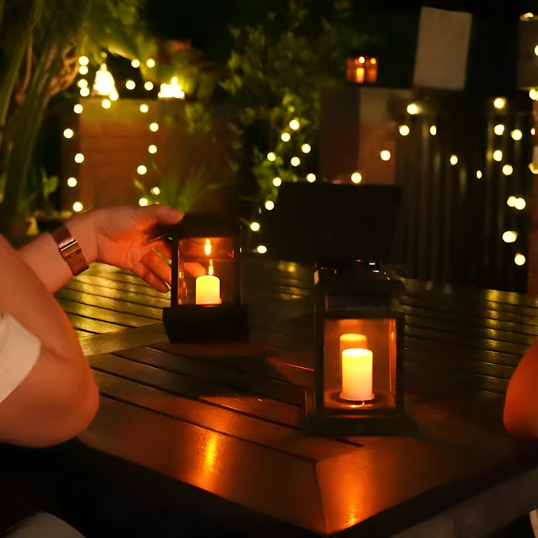 Zwei Laternen mit Kerzen auf einem Holztisch. Hände einer Person sind in der Nähe. Einige Lichter im Hintergrund.