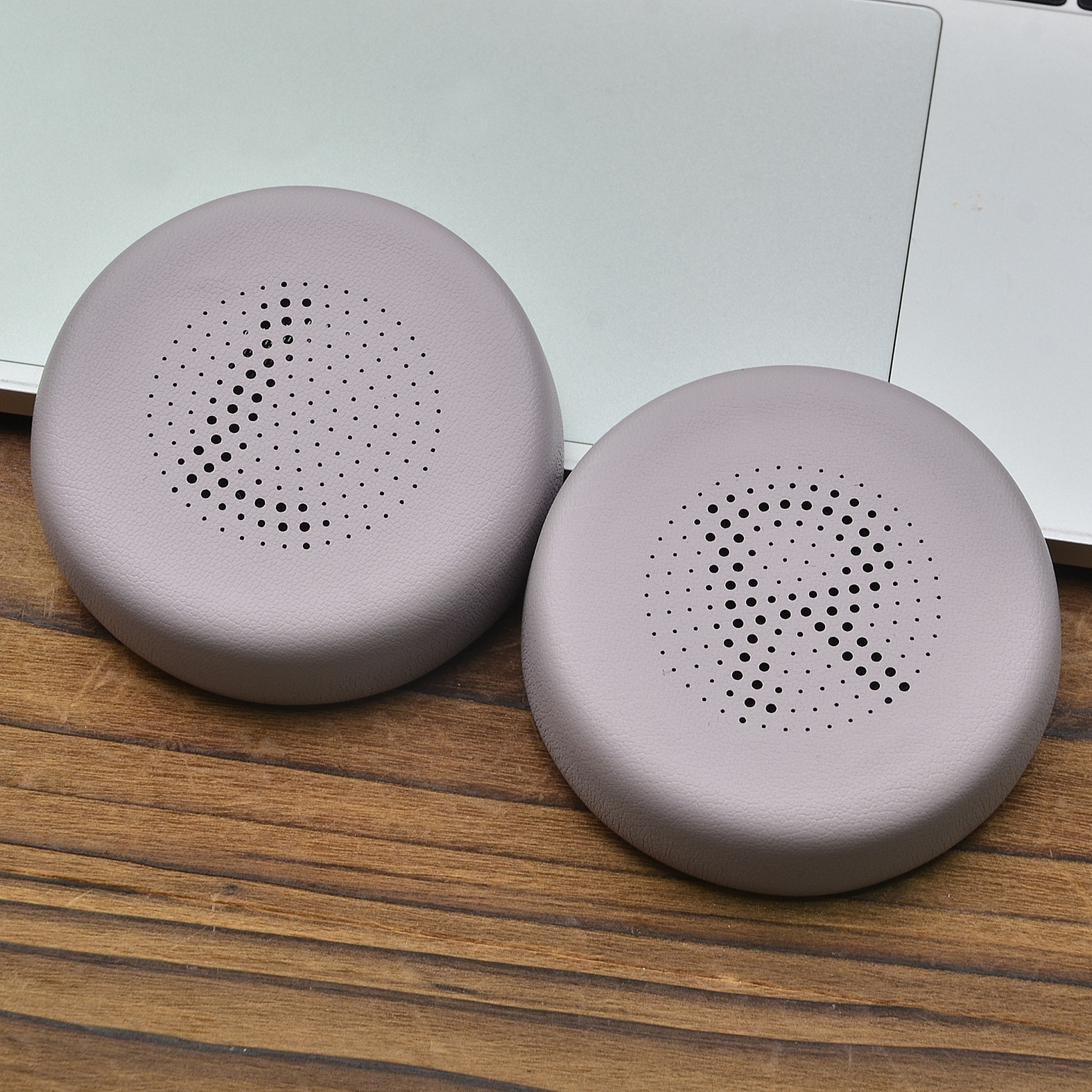 Due cuscinetti auricolari grigi per cuffie con cerchi interni perforati, posizionati su superficie in legno e laptop.