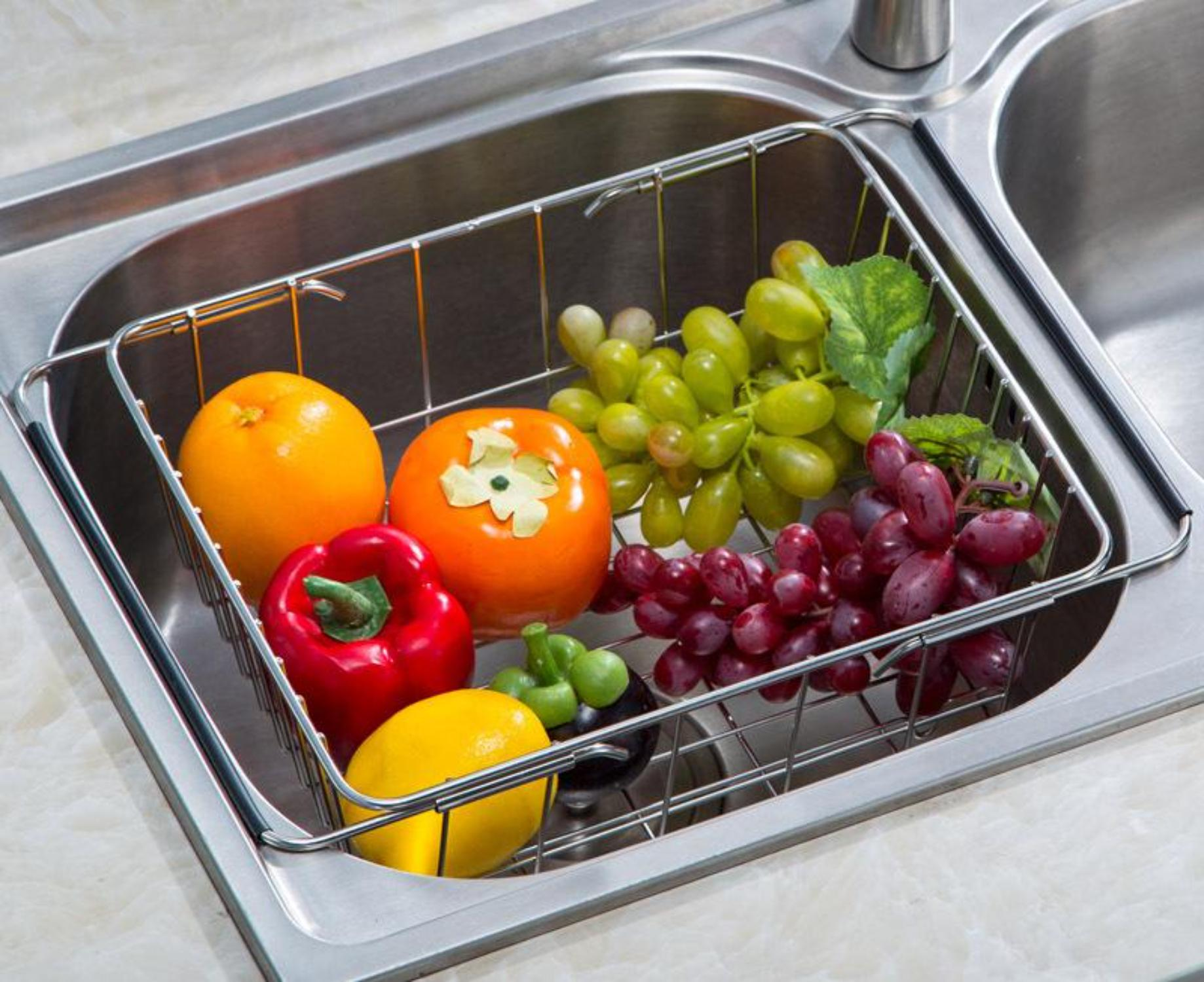 Ein Metallkorb in einem Waschbecken enthält verschiedene Obst- und Gemüsesorten, darunter Orangen, Trauben und Paprika.