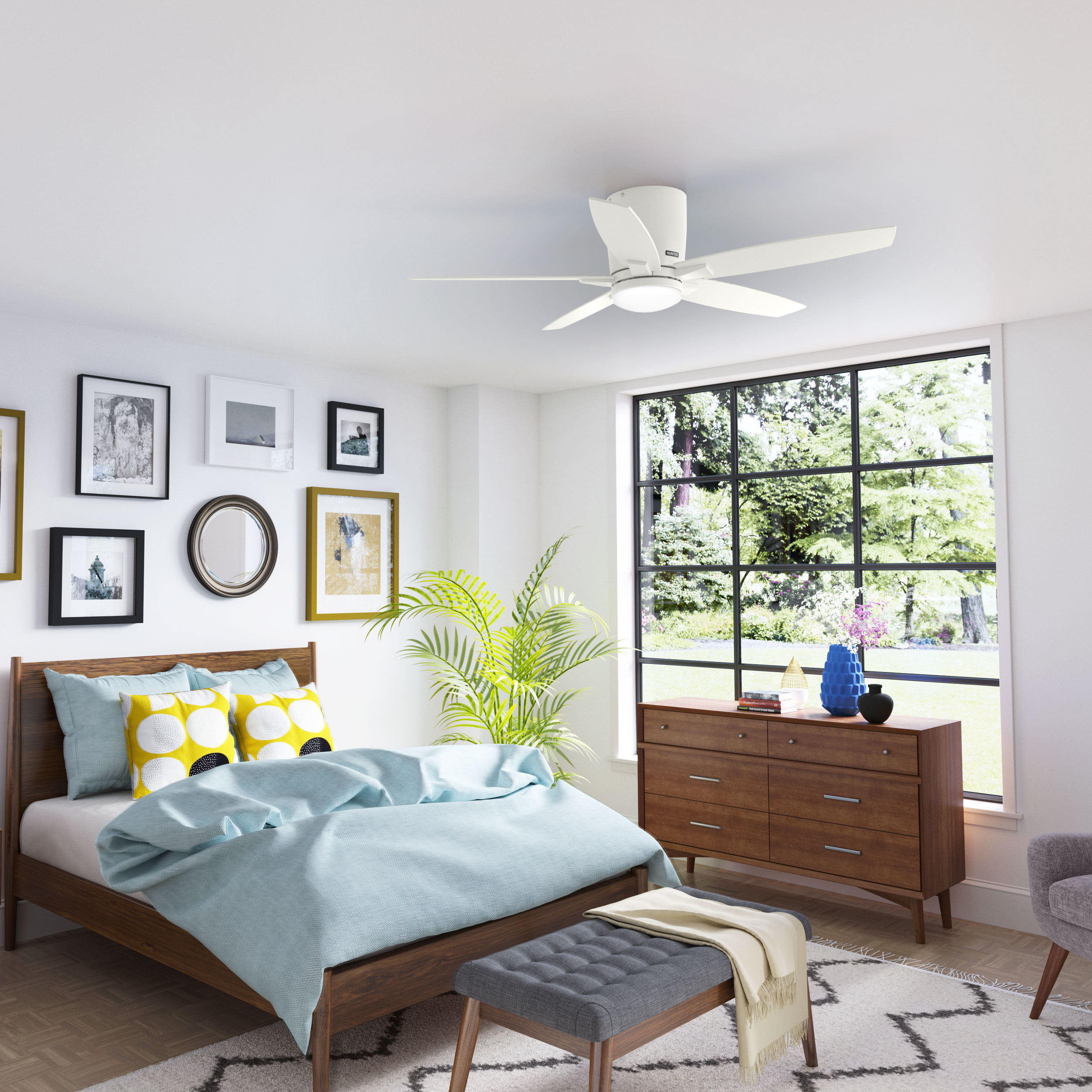 Schlafzimmer mit Bett, Kommode, Kunstwerken und weißem Deckenventilator. Hellblaue Bettwäsche und eine graue Bank.