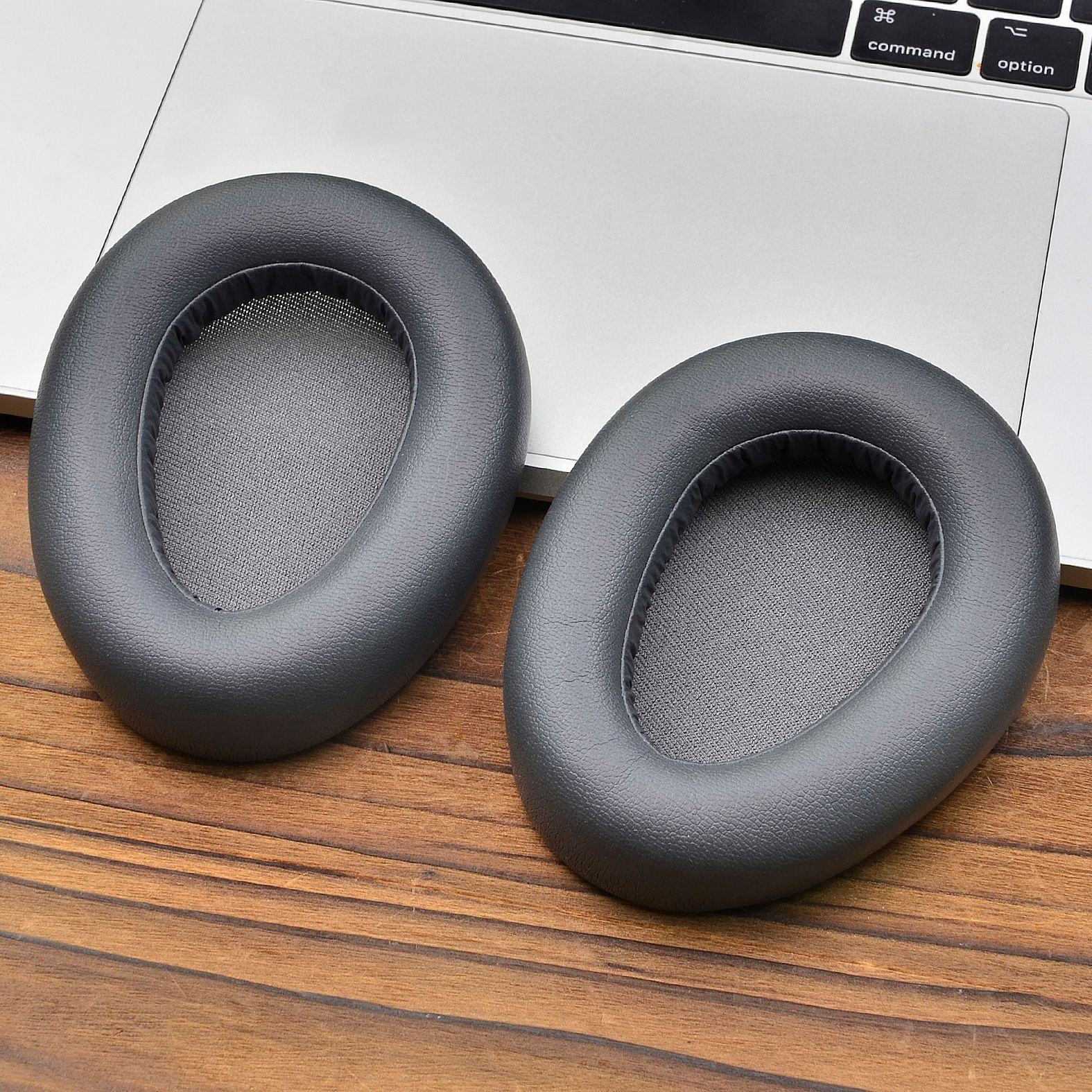 Due cuscinetti auricolari grigi per cuffie su una superficie di legno, probabilmente una scrivania o un tavolo, con un laptop sullo sfondo.