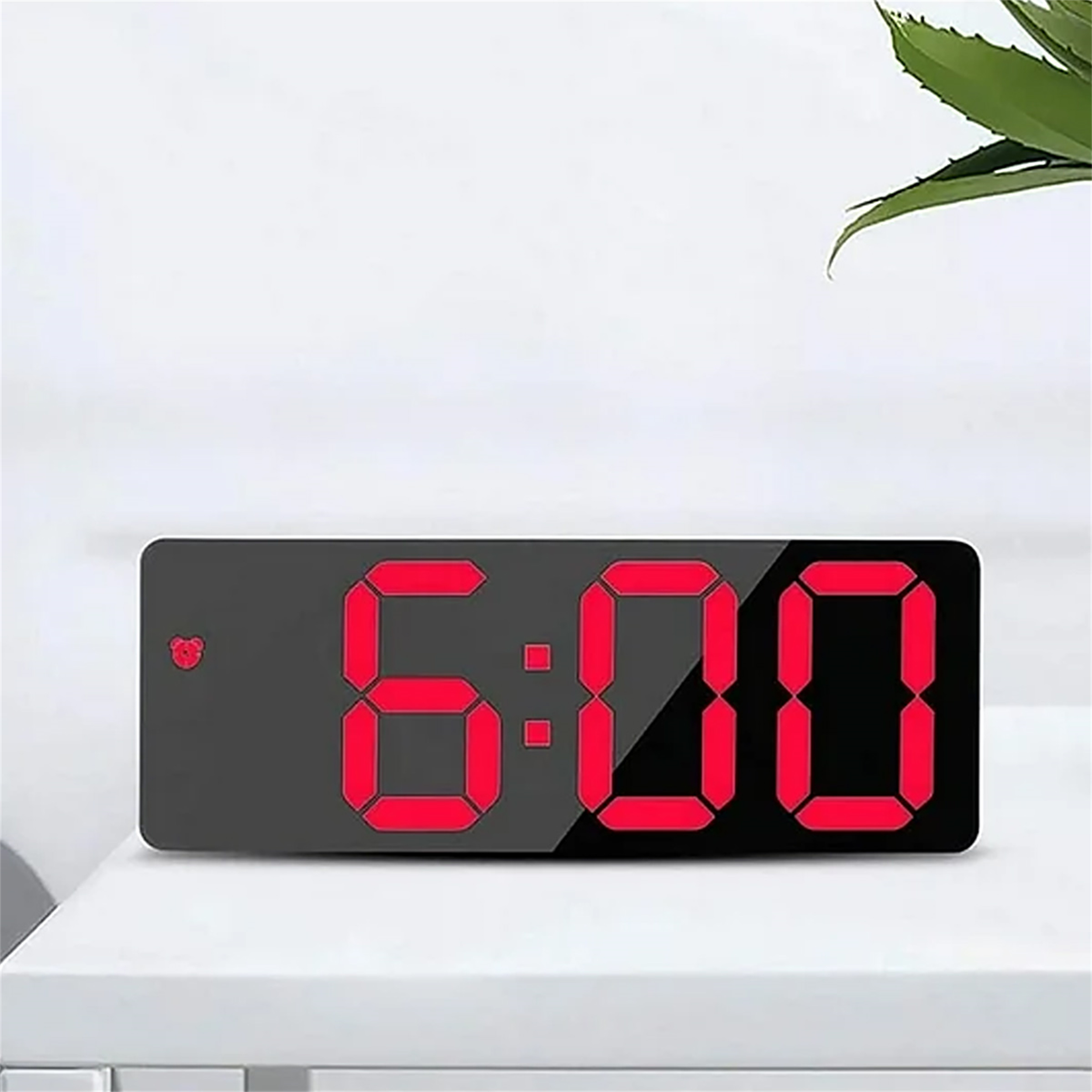 Eine Digitaluhr, die 6:00 in roten Zahlen anzeigt. Sie steht auf einer weißen Oberfläche mit einer Pflanze im Hintergrund.