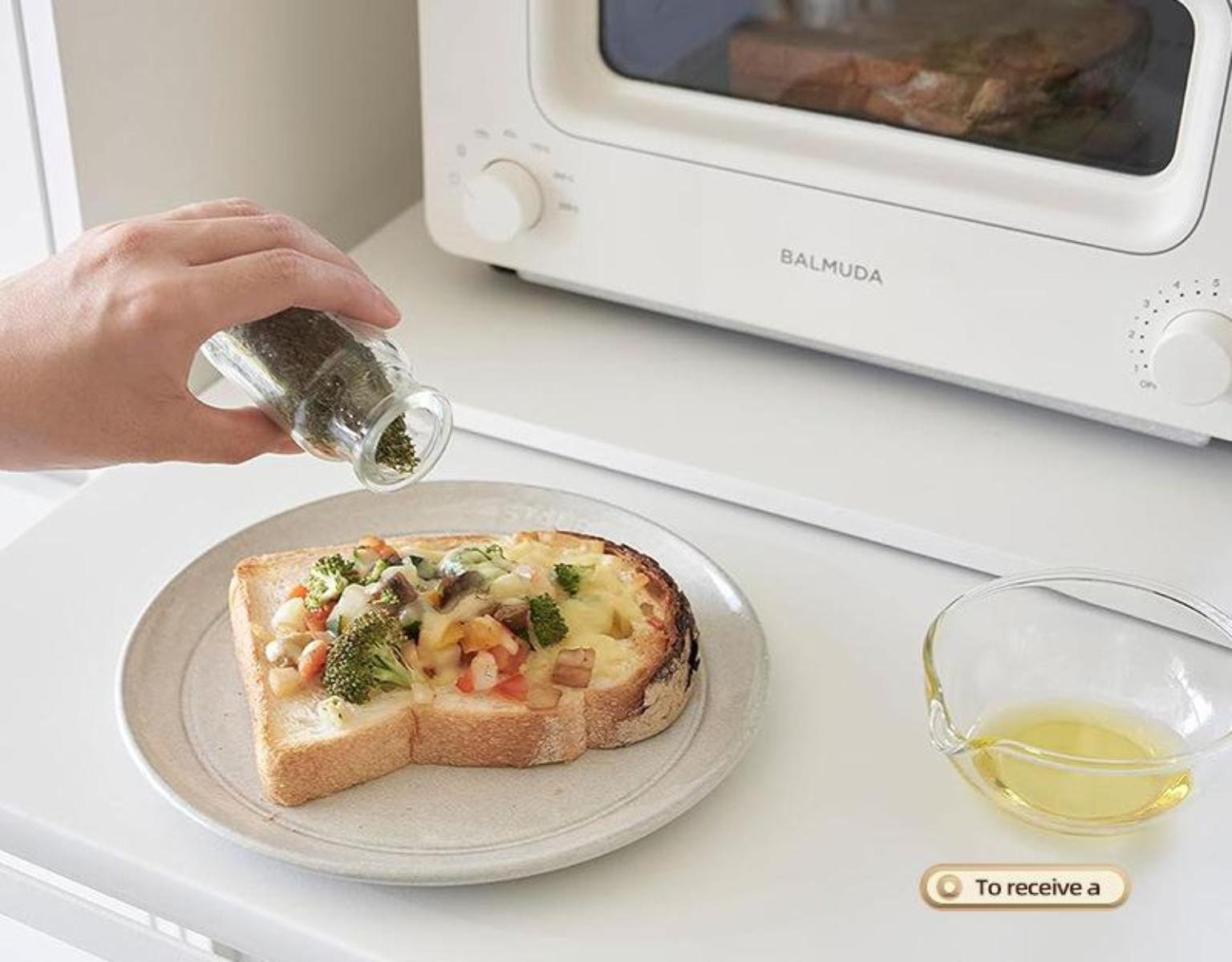 Eine Person bestreut Kräuter auf eine Scheibe Toastbrot mit Belag, neben einem Toaster.