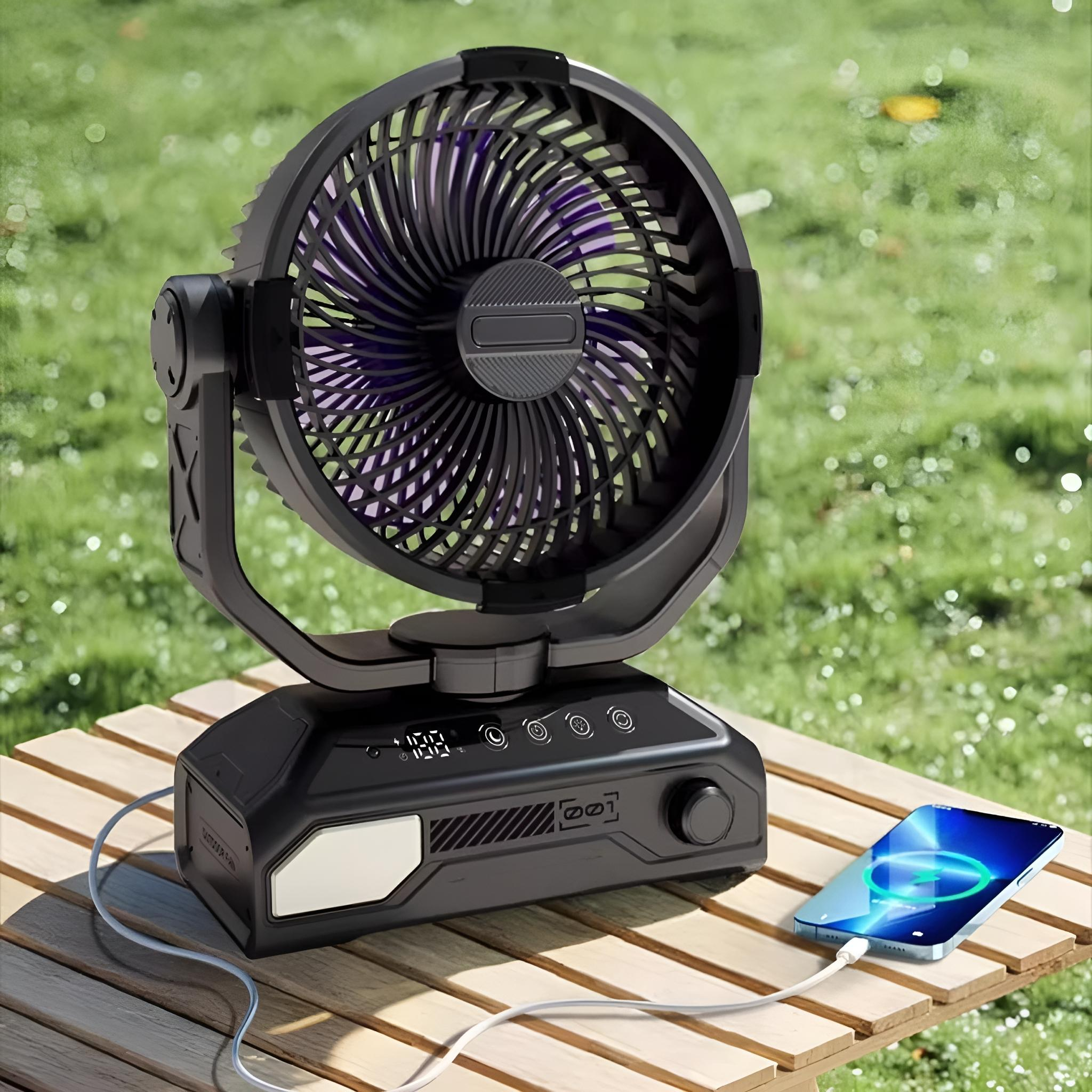 Ein schwarzer Ventilator auf einem Holztisch mit einem ladenden Telefon. Grünes Gras im Hintergrund.