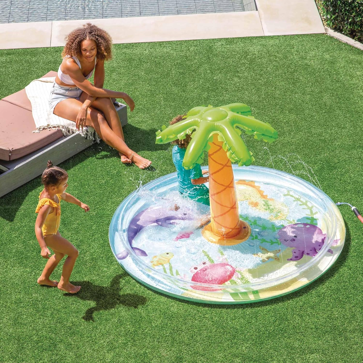 Kinder spielen in einem Planschbecken mit einem Palmen-Wasserfontänen. Eine Frau sitzt auf einer Liege in der Nähe.