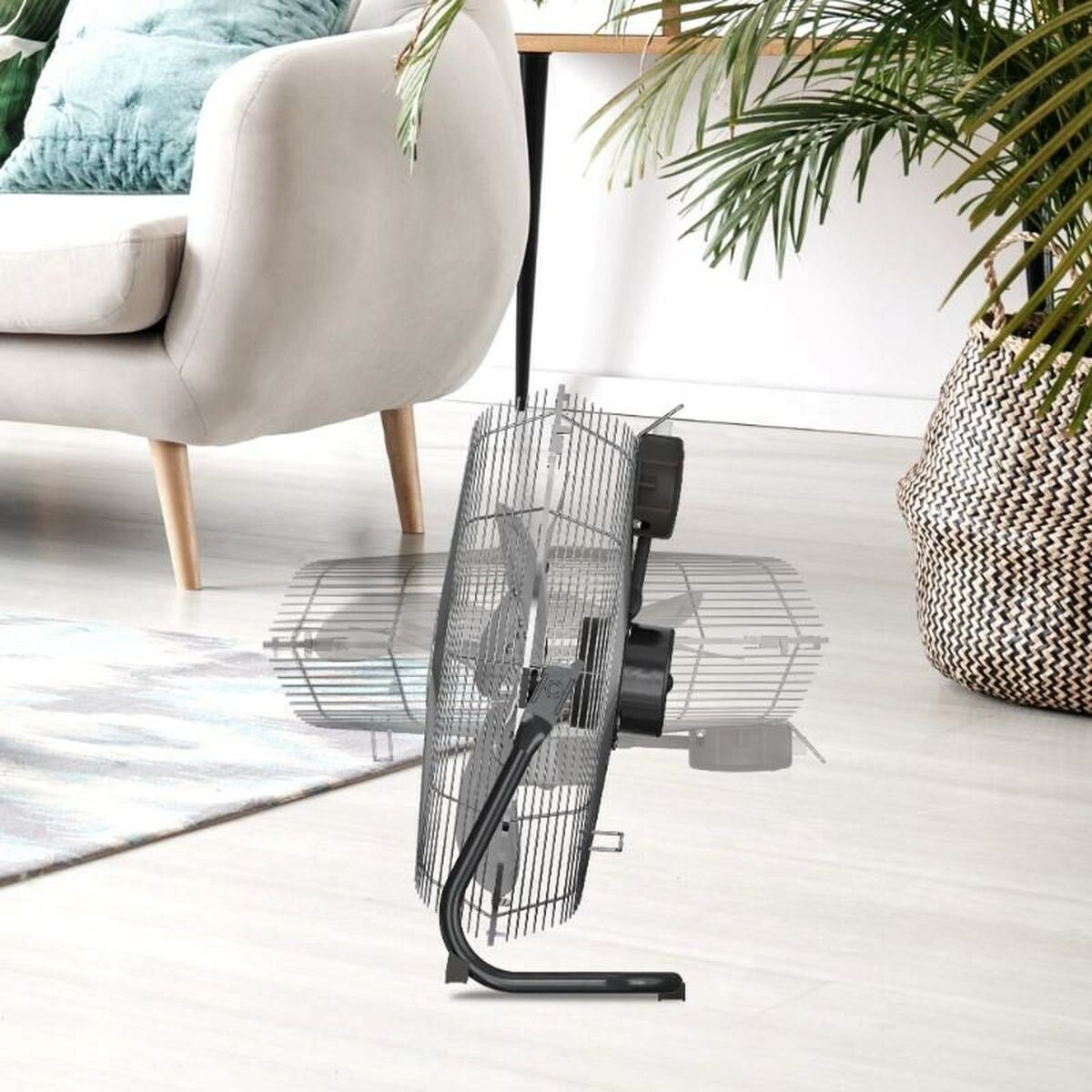 Wentylator elektryczny przechylony w pokoju. Wentylator srebrno-czarny. Tło: kanapa, roślina, drewniana podłoga.