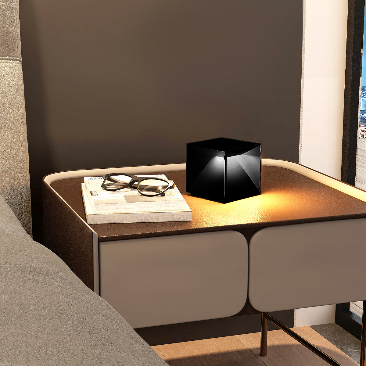 Eine schwarze, würfelförmige Lampe auf einem Nachttisch, neben einem Buch und einer Brille, im Hintergrund ein Fenster.
