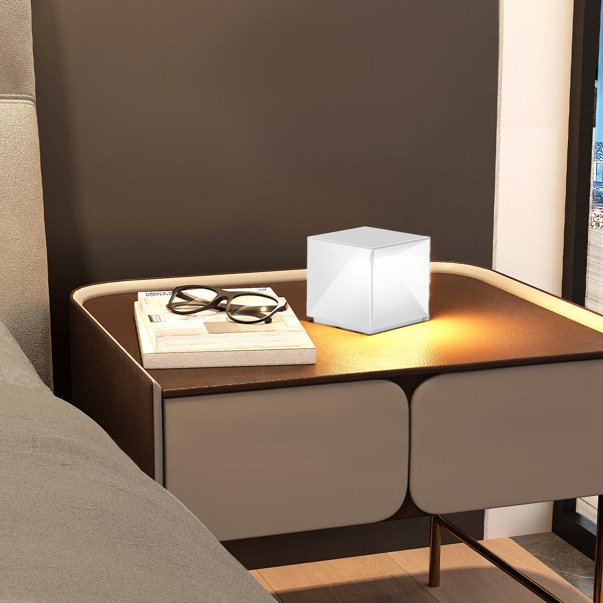 Weißer würfelförmiger Nachttisch mit Lampe, neben einem Bett.