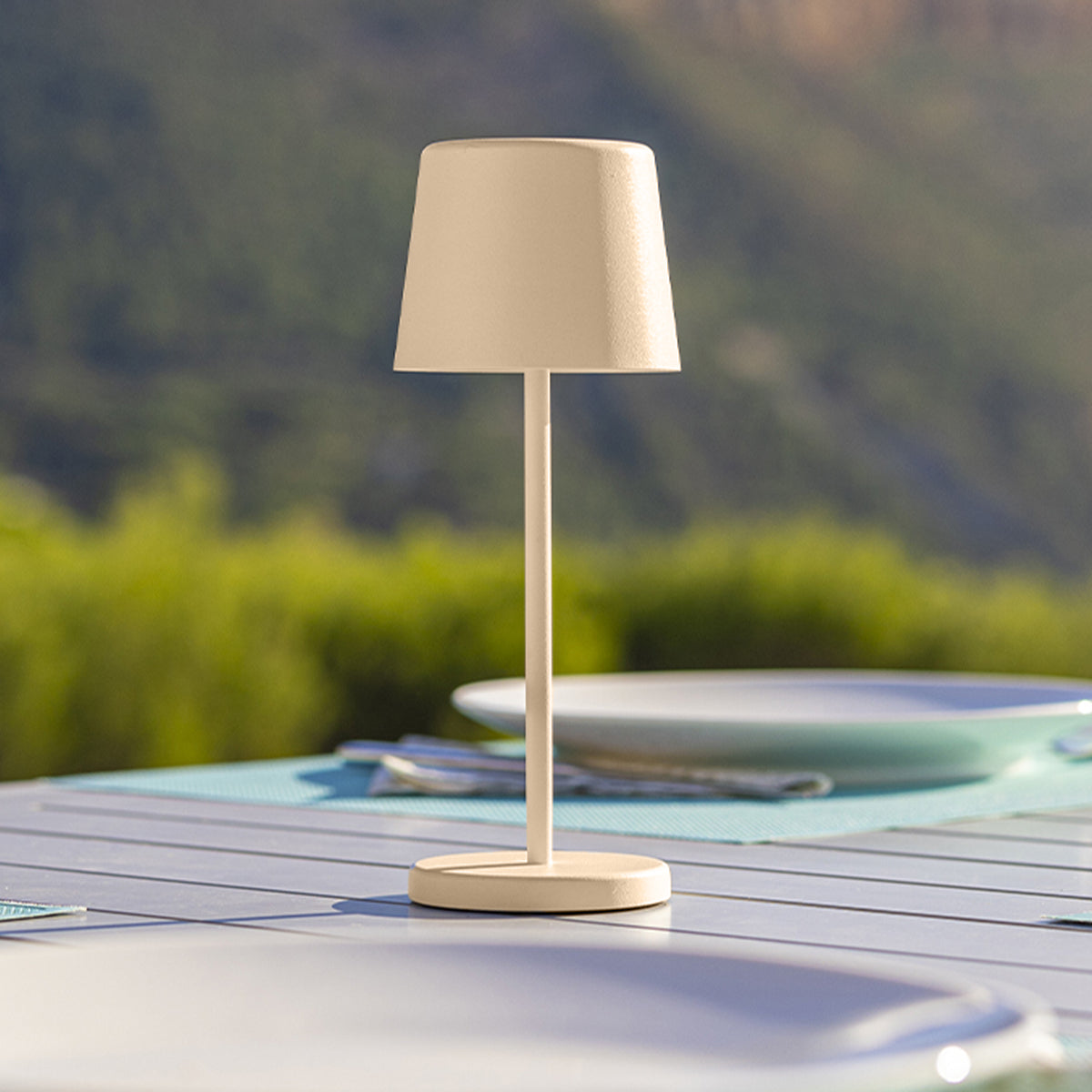 Eine beige Outdoor-Lampe mit konischem Lampenschirm, auf einem Tisch an einem sonnigen Tag, mit grünem Hintergrund.