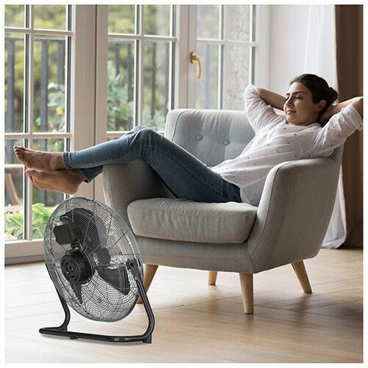 Kobieta relaksuje się w fotelu z wentylatorem na pierwszym planie, w jasnym pokoju.