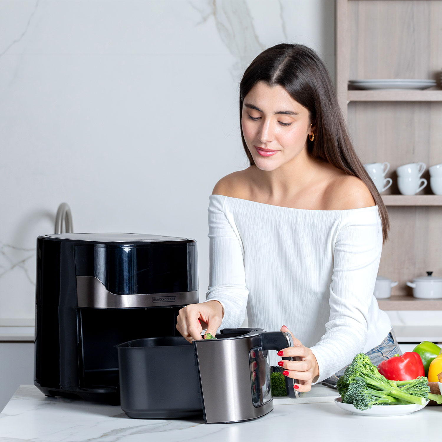Kobieta w kuchni wkłada brokuły do czarnej frytkownicy powietrznej. Warzywa i przybory kuchenne na blacie.