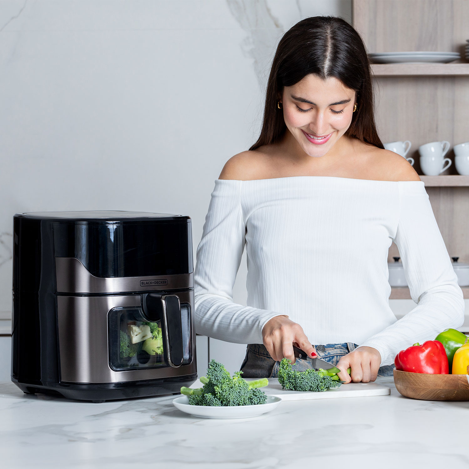 Uśmiechnięta kobieta w bieli kroi brokuły na desce do krojenia obok frytkownicy Black+Decker.