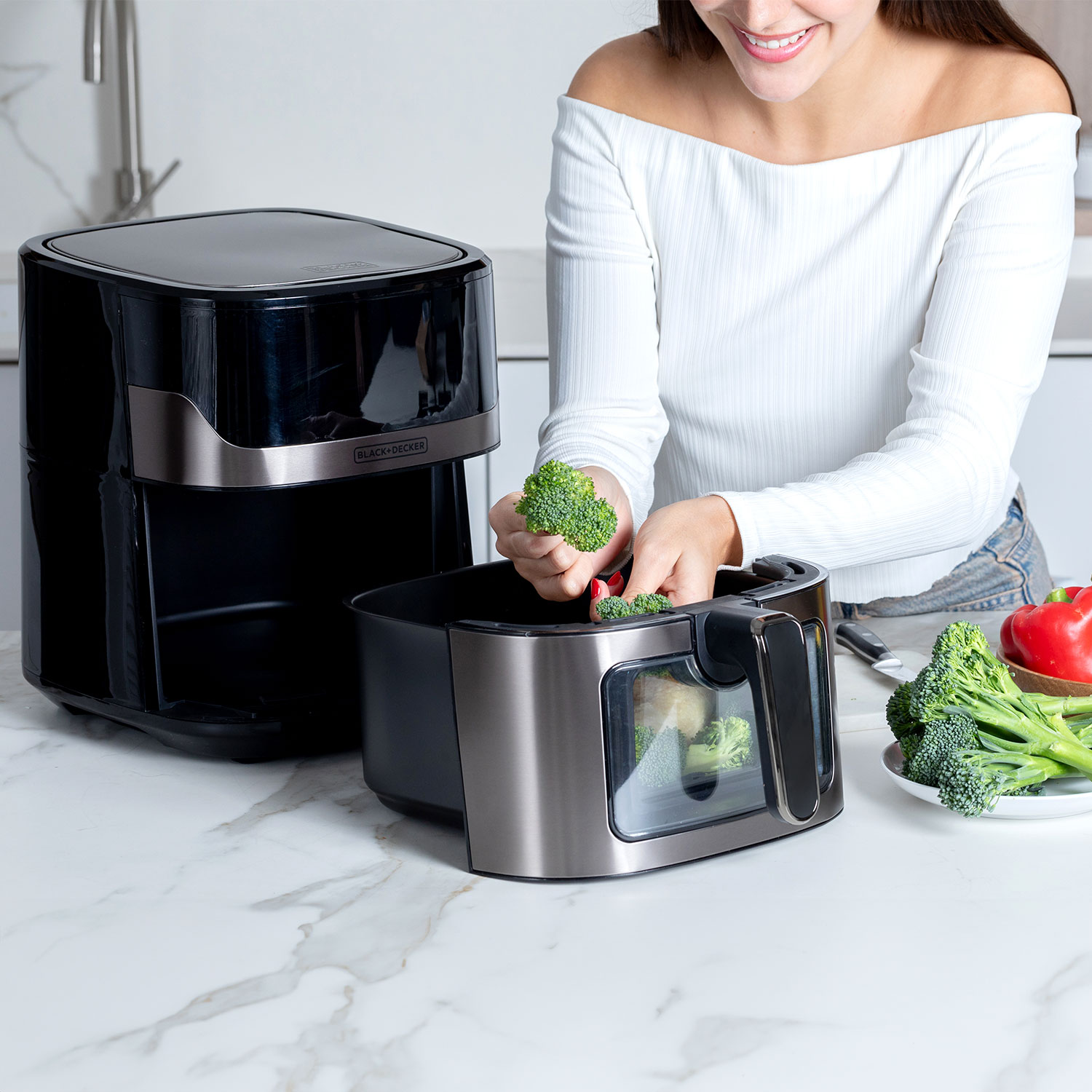 Kobieta wkłada brokuły do czarnej frytkownicy powietrznej. Brokuły, czerwona papryka i inne warzywa na blacie.