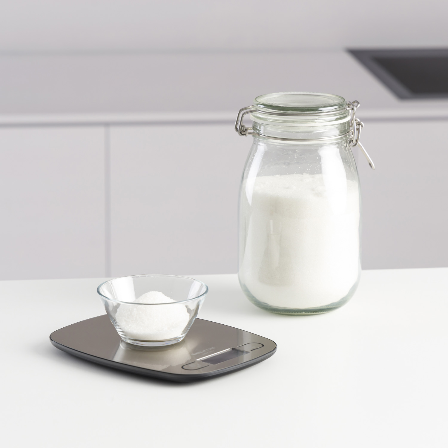 Eine Glasschale und ein Zuckergas stehen auf einer Waage vor einem weißen Küchenhintergrund.