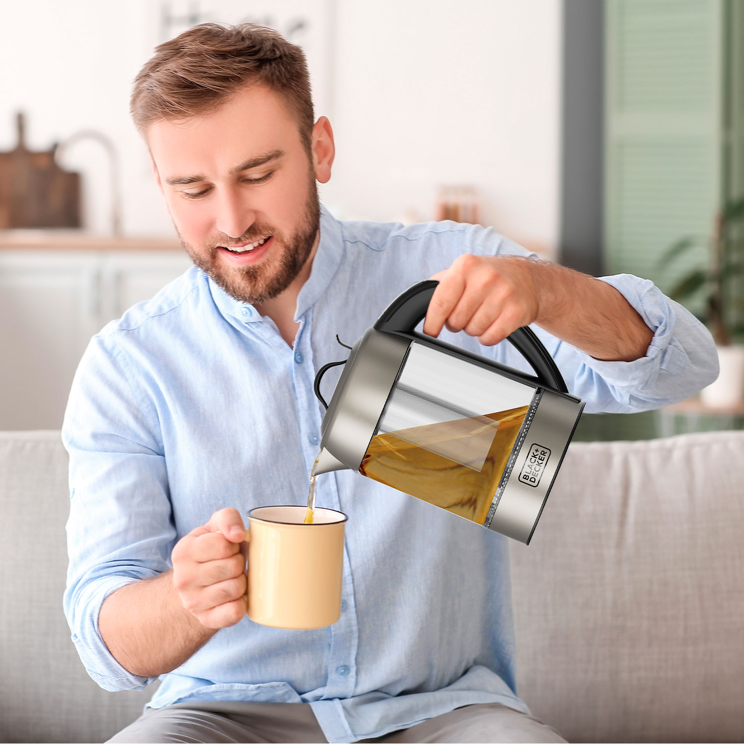 Uśmiechnięty mężczyzna nalewa herbatę z szklanego czajnika do żółtego kubka na szarej sofie.