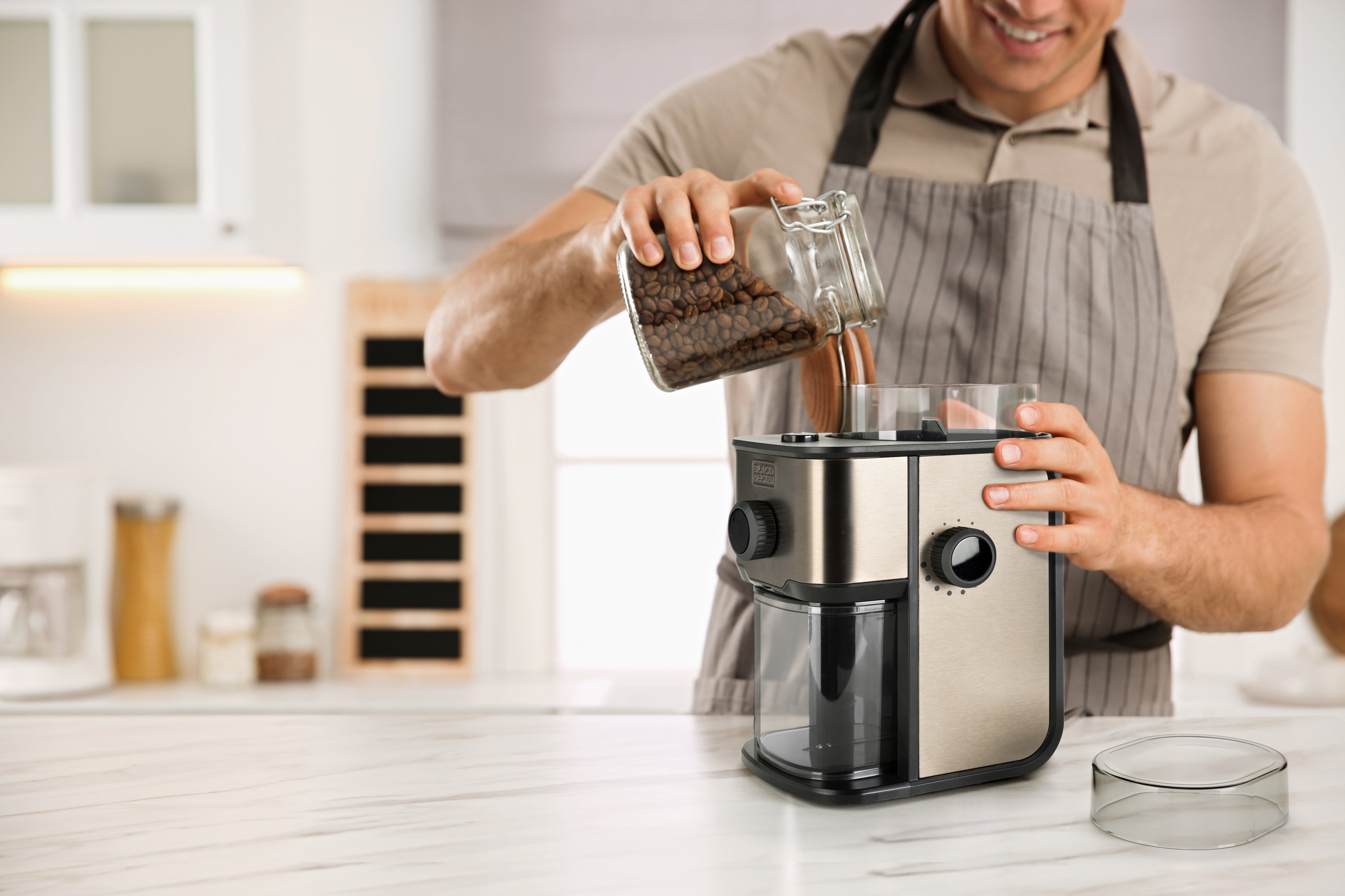 Mężczyzna w fartuchu wsypuje ziarna kawy do srebrnego i czarnego młynka do kawy w białej kuchni.