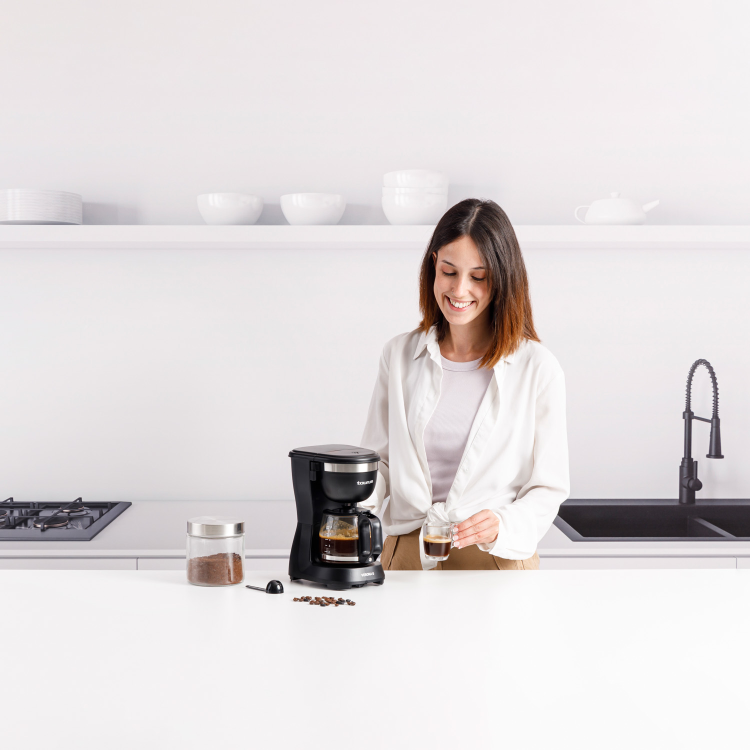 Kobieta w białej koszuli uśmiecha się, trzymając szklankę kawy w jasnej kuchni.