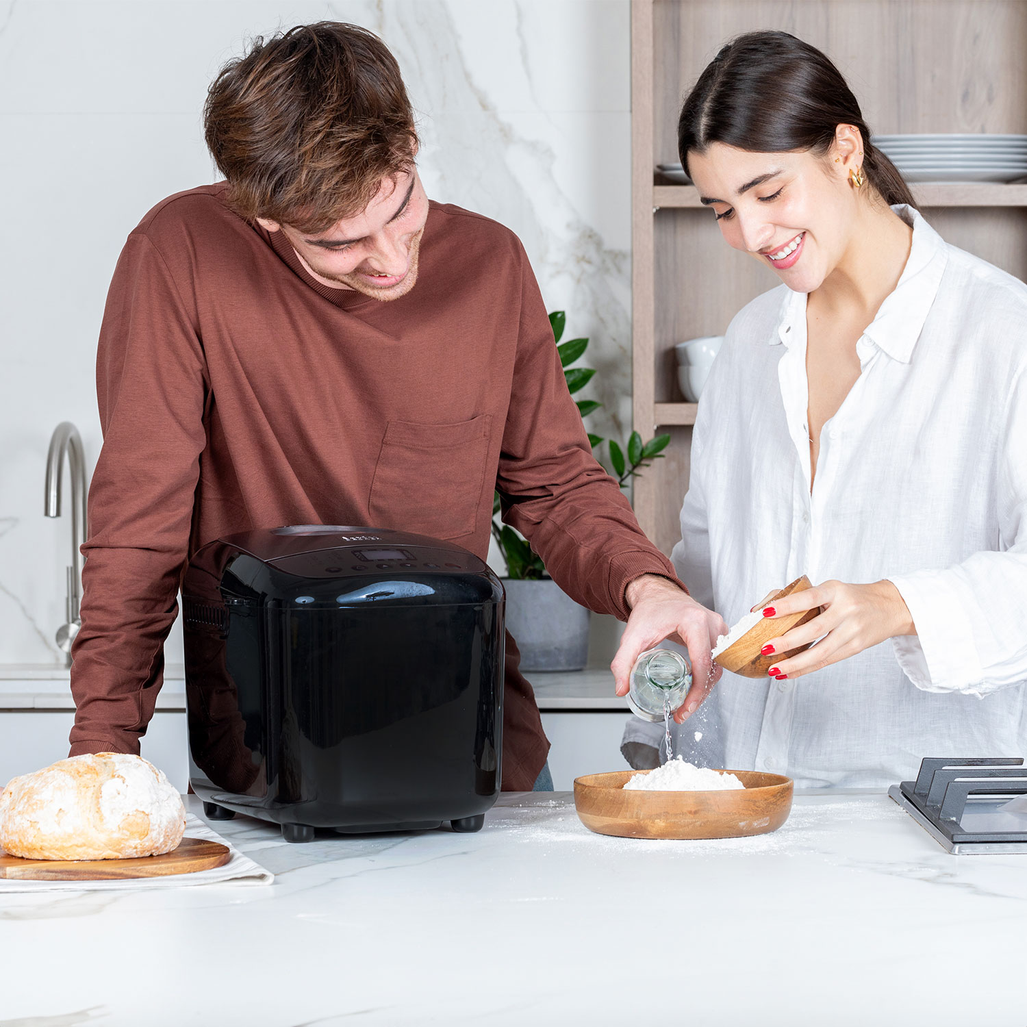 Man kijkt toe terwijl vrouw ingrediënten in een houten kom giet naast een zwarte broodbakmachine op een wit aanrecht.