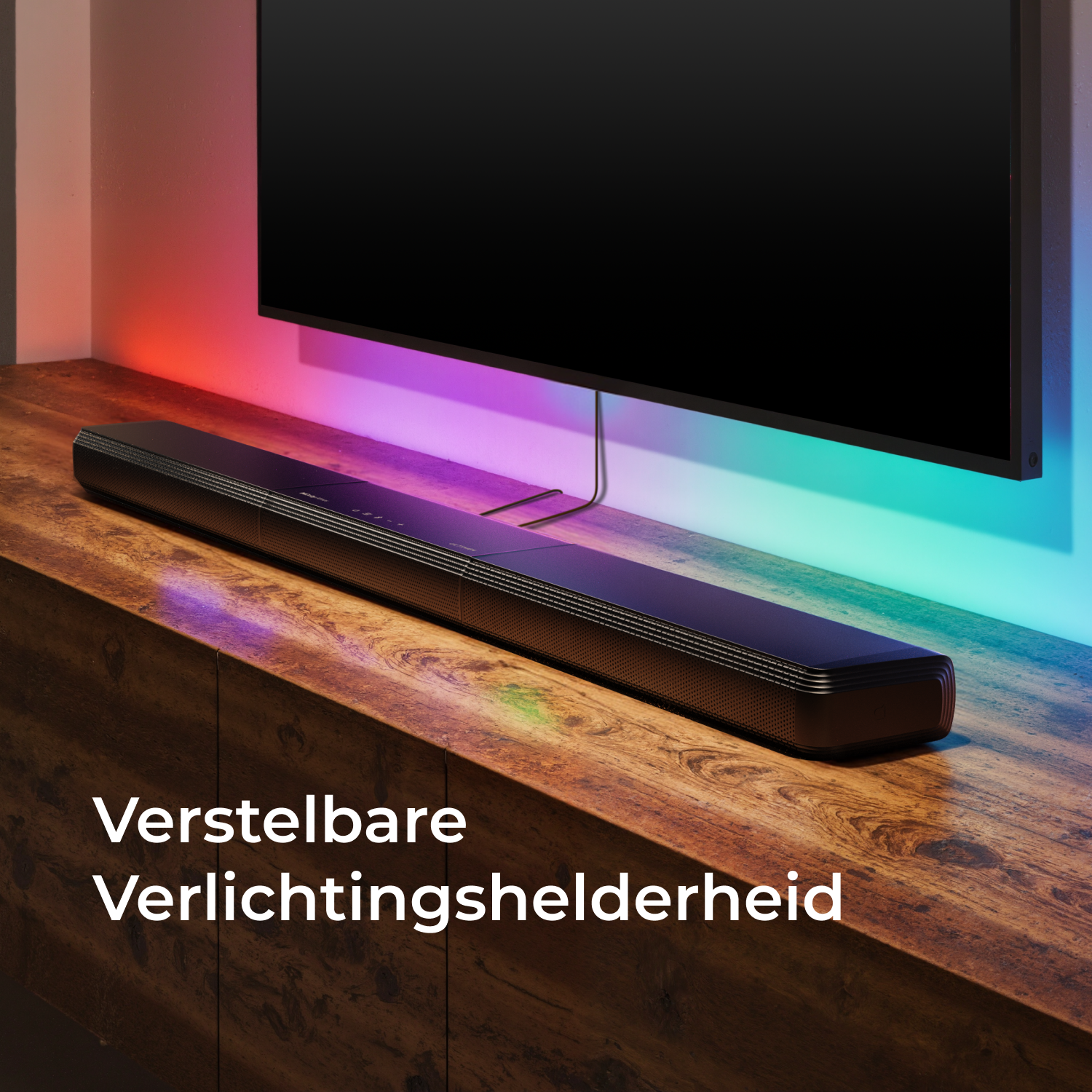A black soundbar placed on a wooden surface below a TV, with adjustable lighting effects on the wall above.
