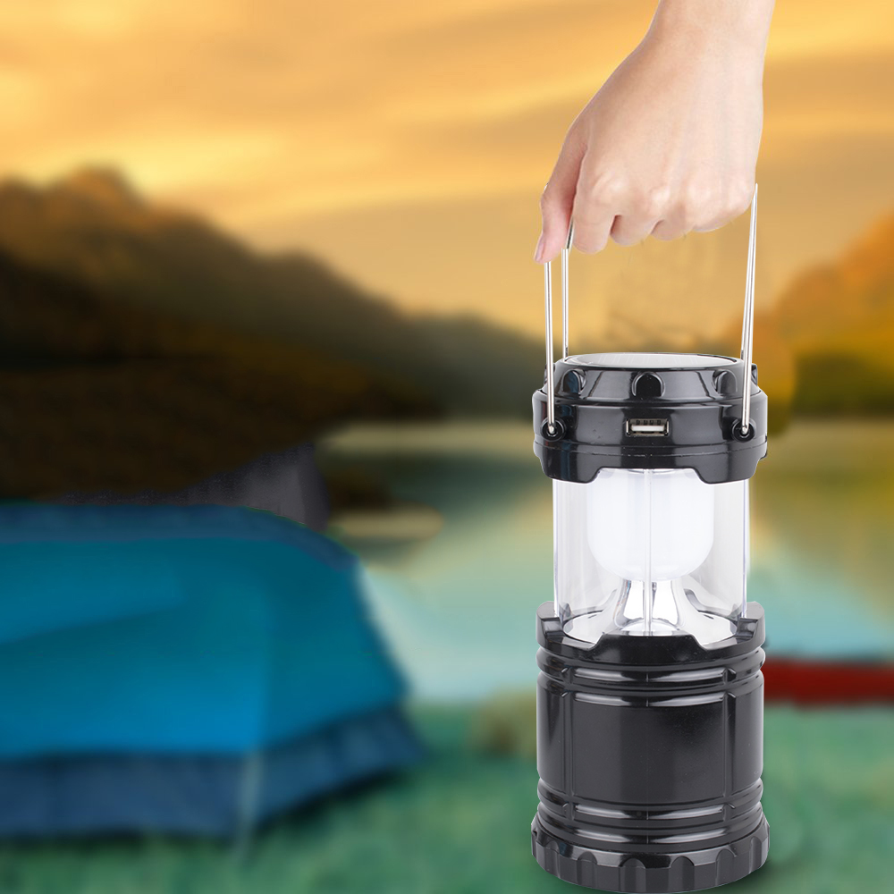 Eine Hand, die eine schwarze Campinglaterne mit weißem Licht hält, im Freien mit Naturhintergrund.