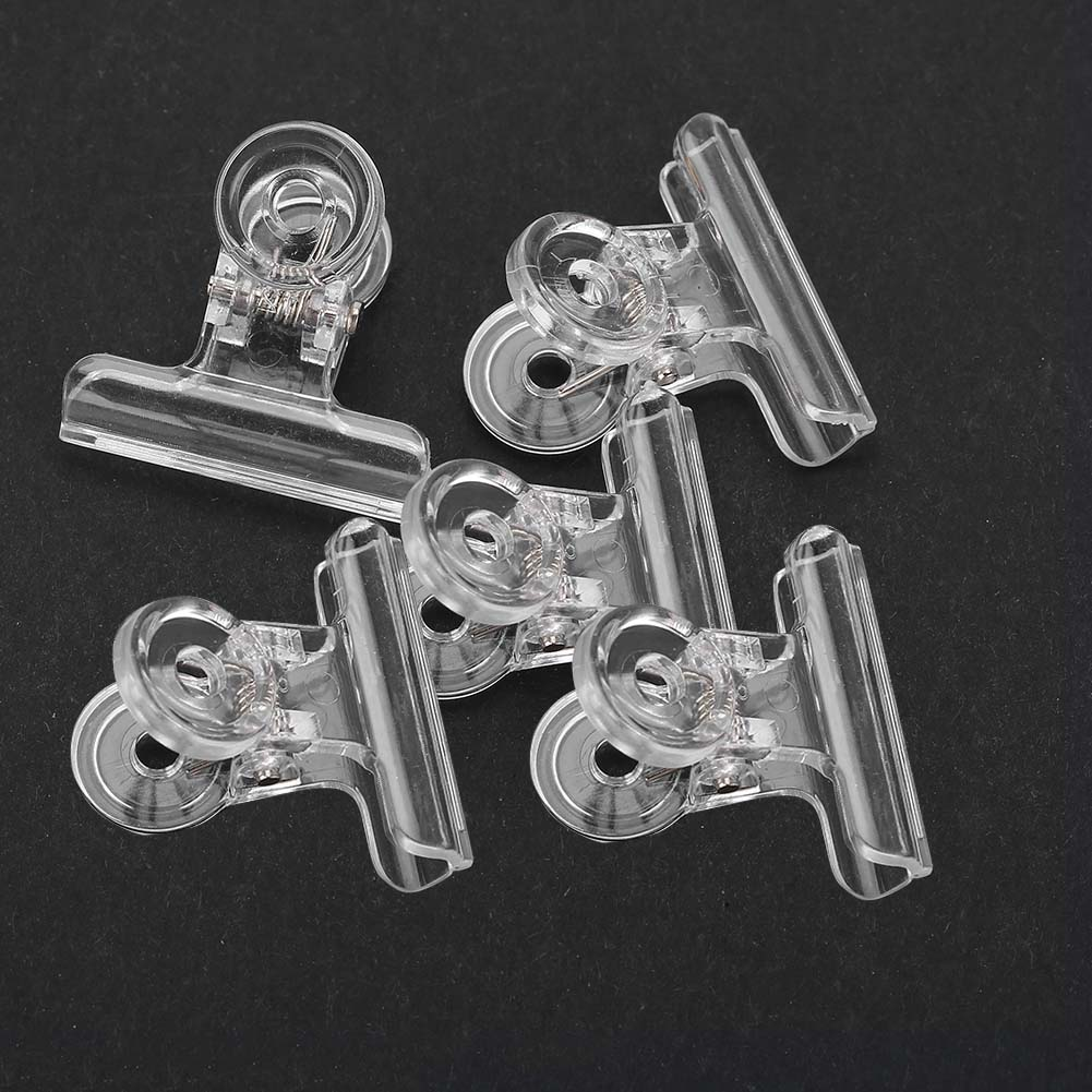 Fünf transparente Nagelkunst-Clips auf dunkler Oberfläche. Jeder Clip hat eine gebogene Oberseite und einen Federmechanismus.