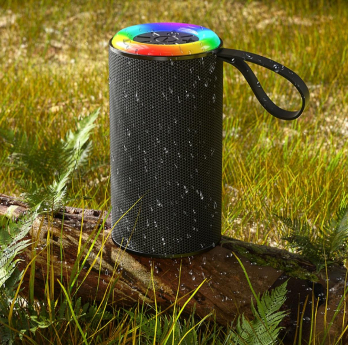Ein schwarzer Lautsprecher mit einem Regenbogen-Lichtring sitzt auf einem Baumstamm mit Gras. Wassertröpfchen sind vorhanden.