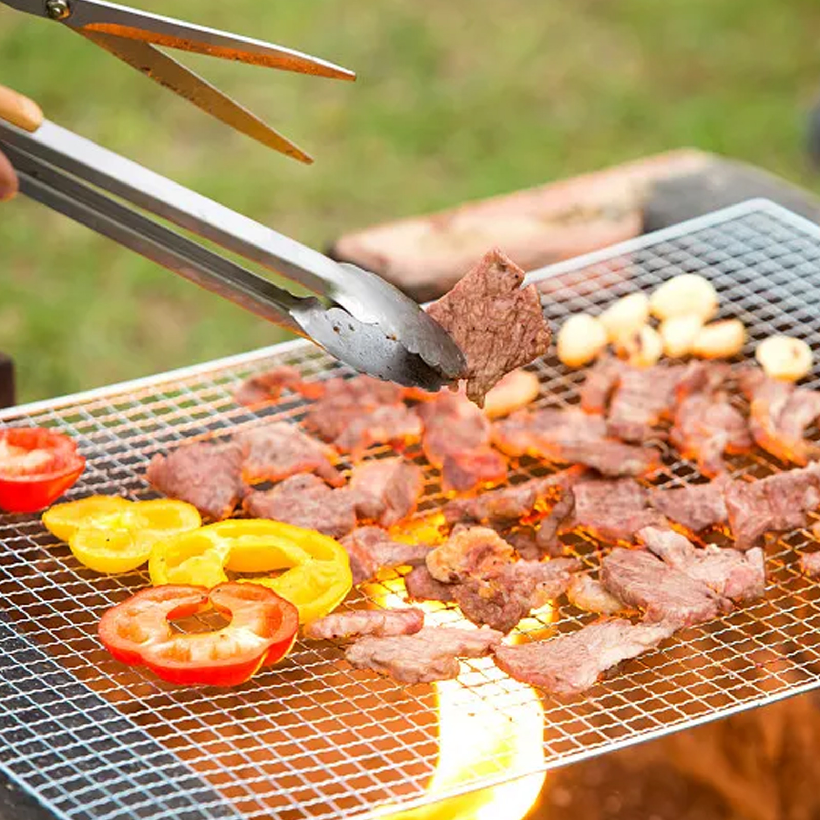 Person verwendet Zangen, um Fleischstücke auf einem Grill zu wenden. Tomaten und gelbe Paprika sind ebenfalls auf dem Grill.