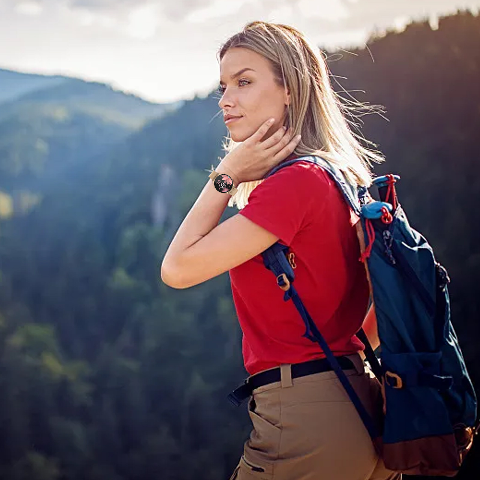 Eine Frau mit blonden Haaren wandert und trägt ein rotes Hemd, khakifarbene Hosen und einen Rucksack; eine Smartwatch am Handgelenk.
