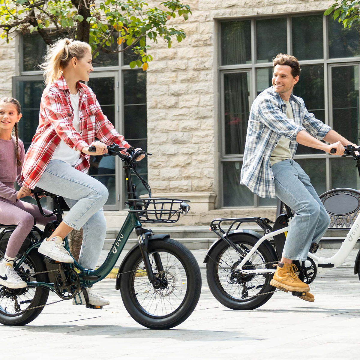 Rodzina jeździ na rowerach po utwardzonej ścieżce. Rowery są zielone i białe. Tło to kamienny budynek.