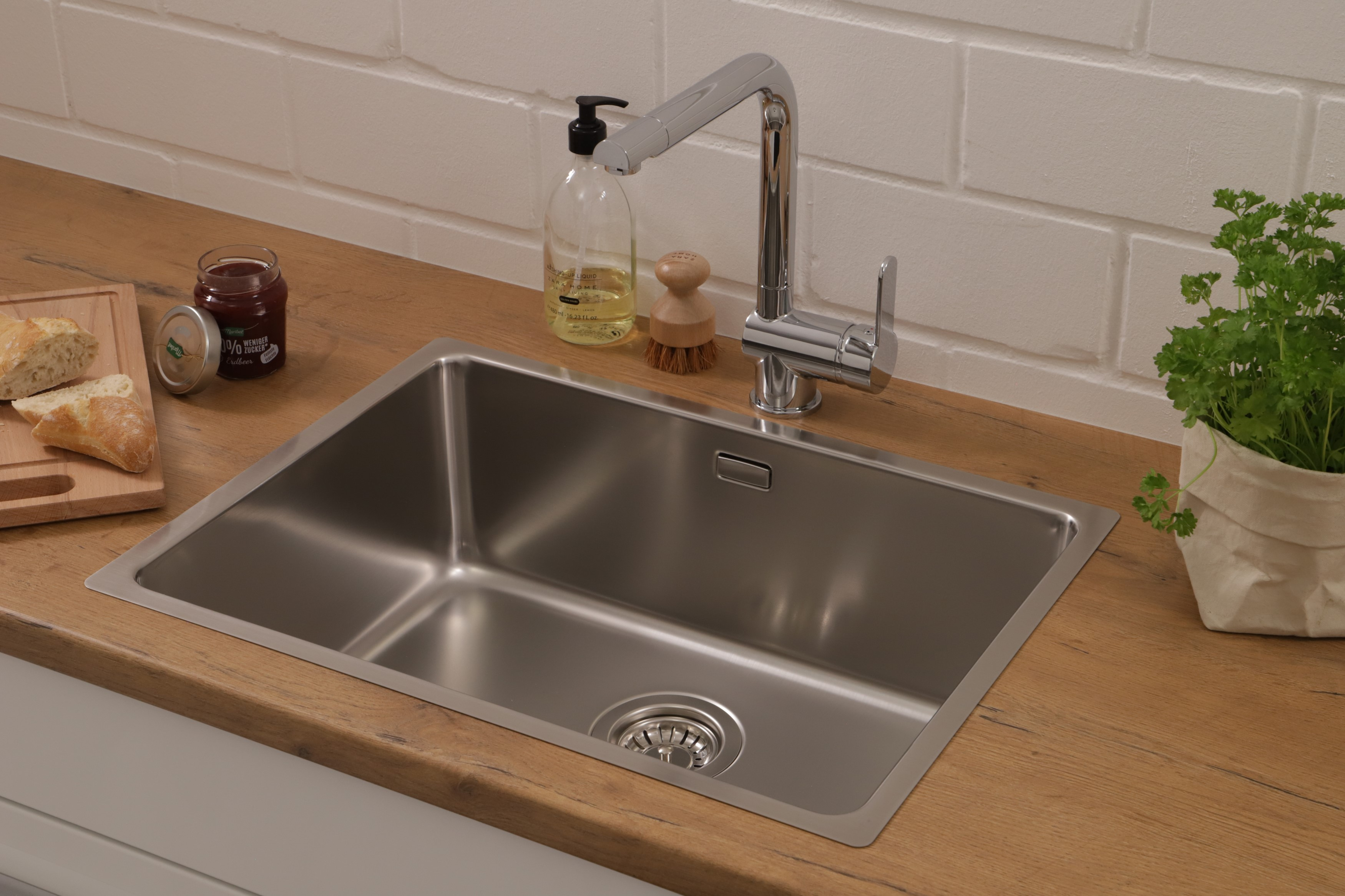 Ein Edelstahlwaschbecken mit Wasserhahn, Seifenspender und Holz-Arbeitsplatte vor einer Ziegelmauer.