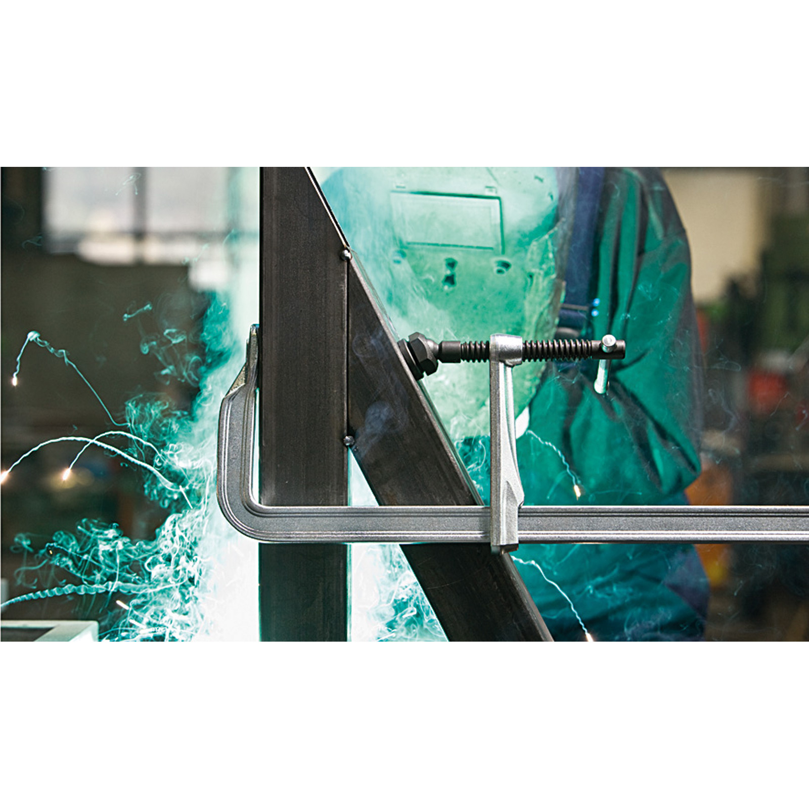 Eine Metallklemme hält zwei Metallträger. Schweißfunken sind sichtbar. Die Szene ist in einer Werkstatt.