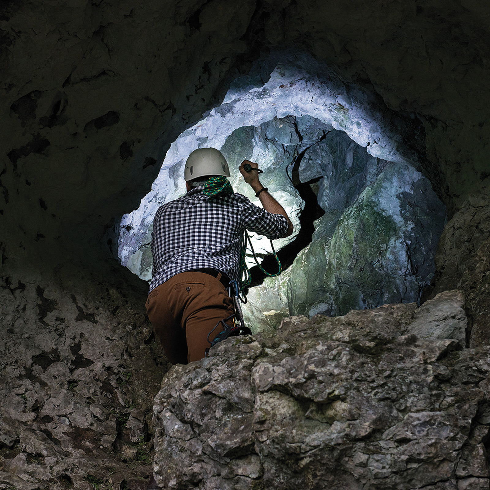 Osoba w jaskini, używająca czołówki, patrzy na oświetlony otwór w skałach.