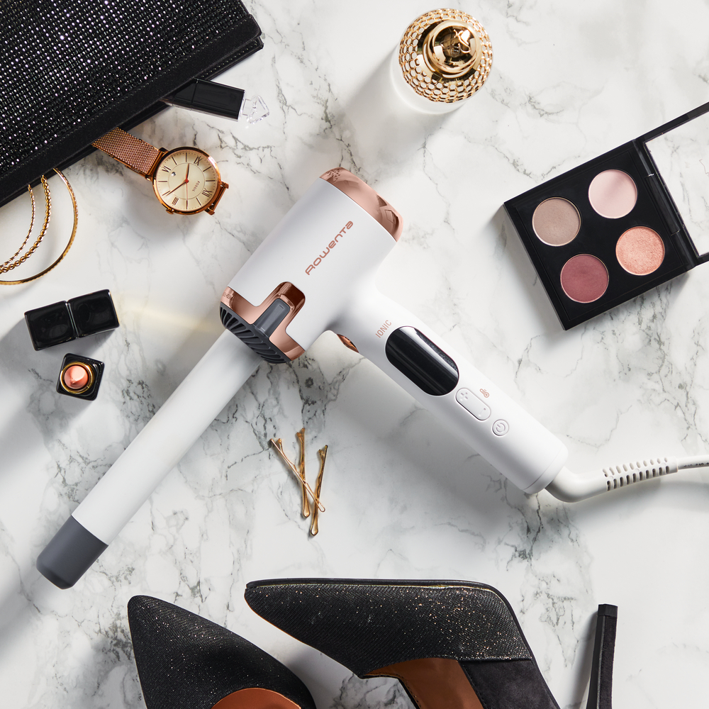 Biała i różowo-złota suszarka do włosów z akcesoriami, takimi jak makijaż, zegarek i szpilki na marmurowej powierzchni.